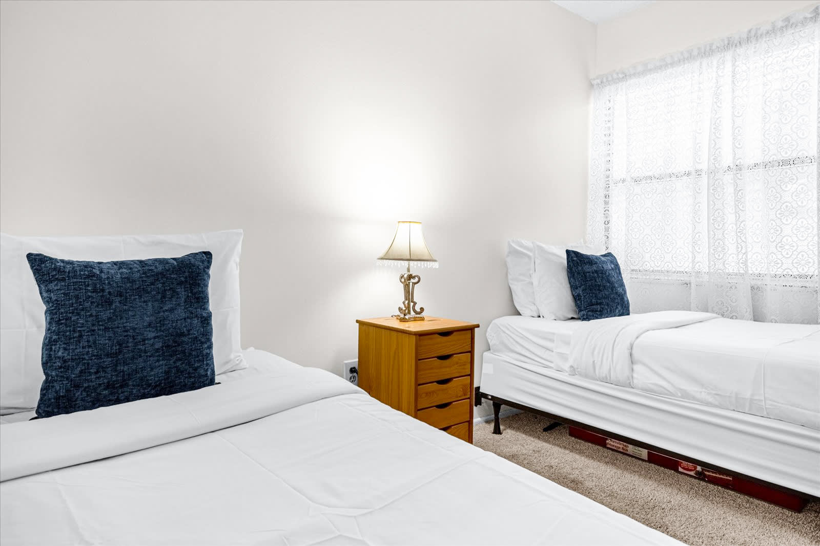 This bedroom offers two white twin beds with blue accent pillows, light beige carpet, and a wooden nightstand with a lamp. A window with sheer white curtains provides soft natural light.