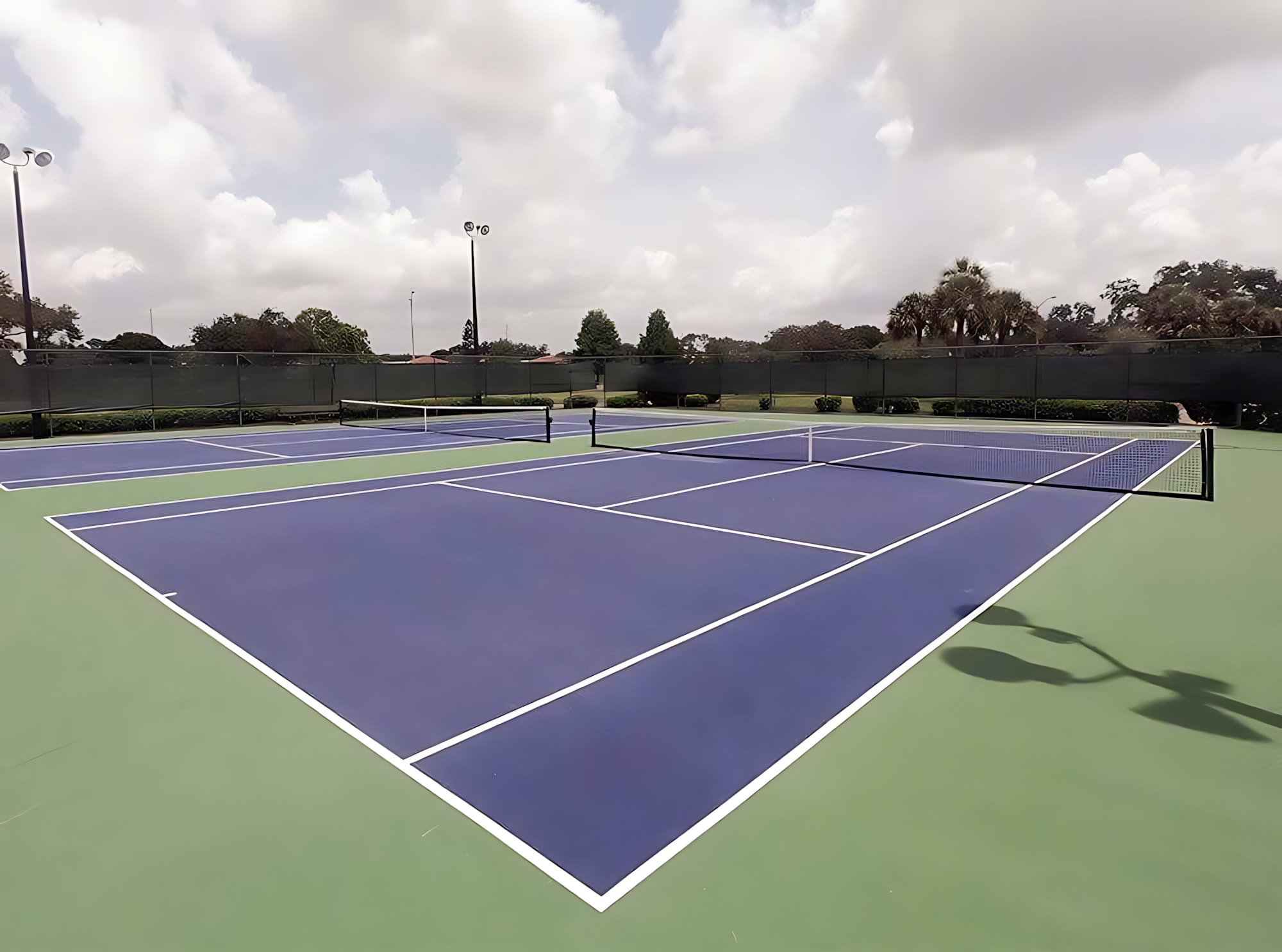 Two side-by-side tennis courts with blue playing surfaces and green surrounds, enclosed by a dark fence. The courts are well-maintained under a partly cloudy sky, with tall light poles for evening play.
