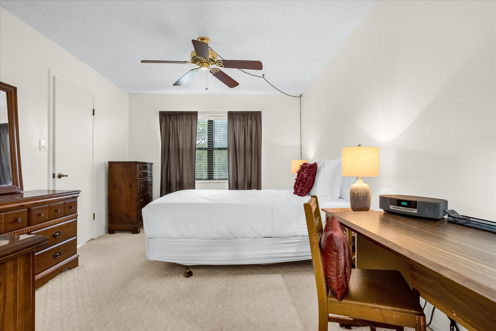 A cozy bedroom with a white queen-sized bed, light beige carpet, and a window framed by dark brown curtains. The room is furnished with a wooden dresser, a nightstand with a lamp, and a wooden desk with a chair.