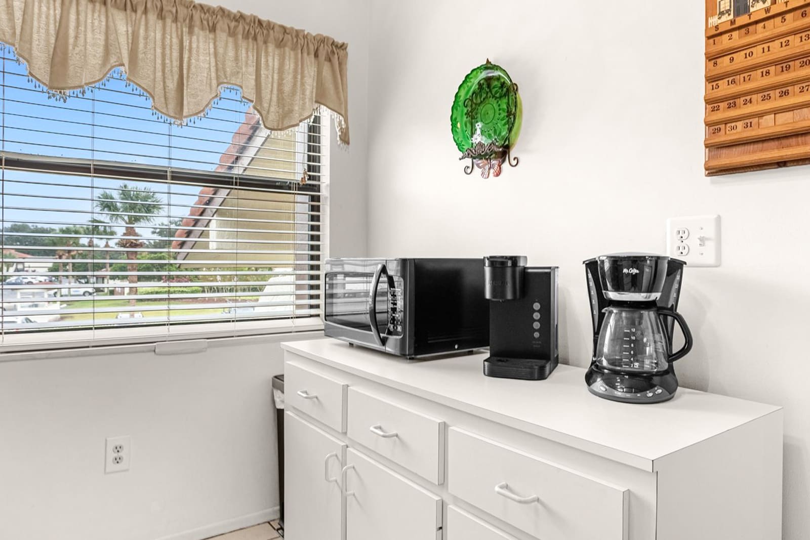 A dedicated counter space with white lower cabinets features a black microwave, a single-serve coffee maker, and a drip coffee maker. A large window with horizontal blinds offers a view of the outdoors.