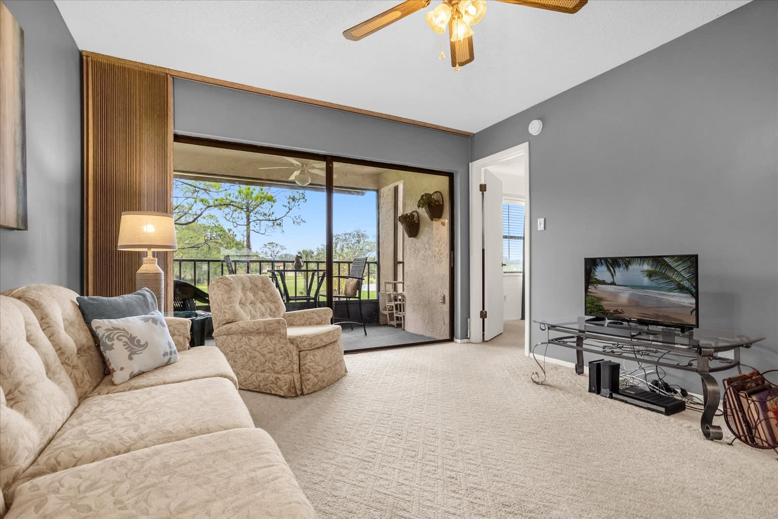 A bright living room features a patterned beige sofa and armchair, light beige carpet, and gray walls, with a ceiling fan and a sliding glass door leading to a balcony overlooking a golf course.