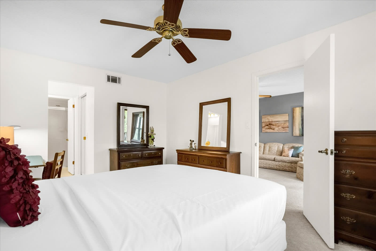 This bedroom features a white queen-sized bed, light beige carpet, and a wooden dresser with a large mirror. A doorway leads to an adjacent living area, and a ceiling fan is mounted overhead.