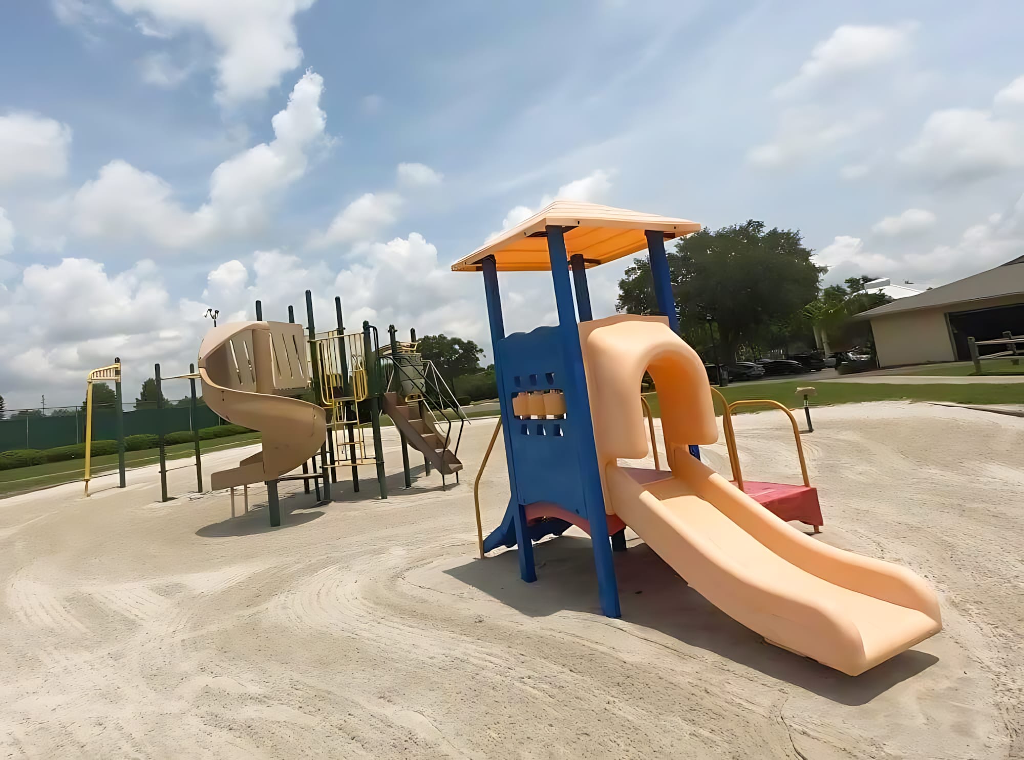 A vibrant outdoor playground with multiple slides, climbing structures, and a sandy ground cover. The play area is set under a bright, partly cloudy sky, surrounded by green landscaping and distant buildings.