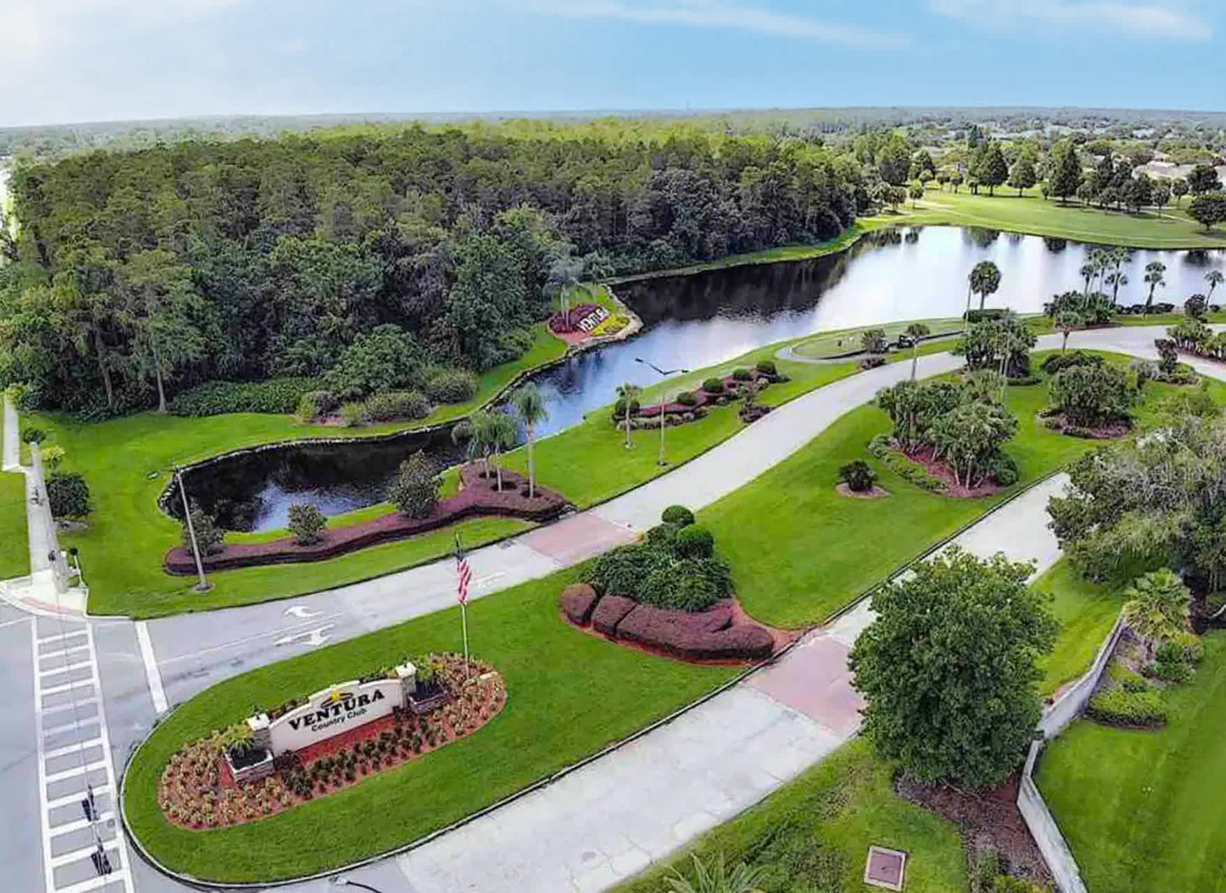 An aerial view showcases the entrance to Ventura Country Club, featuring a manicured landscape with lush green lawns, palm trees, a winding road, and a large lake, all under a bright sky.