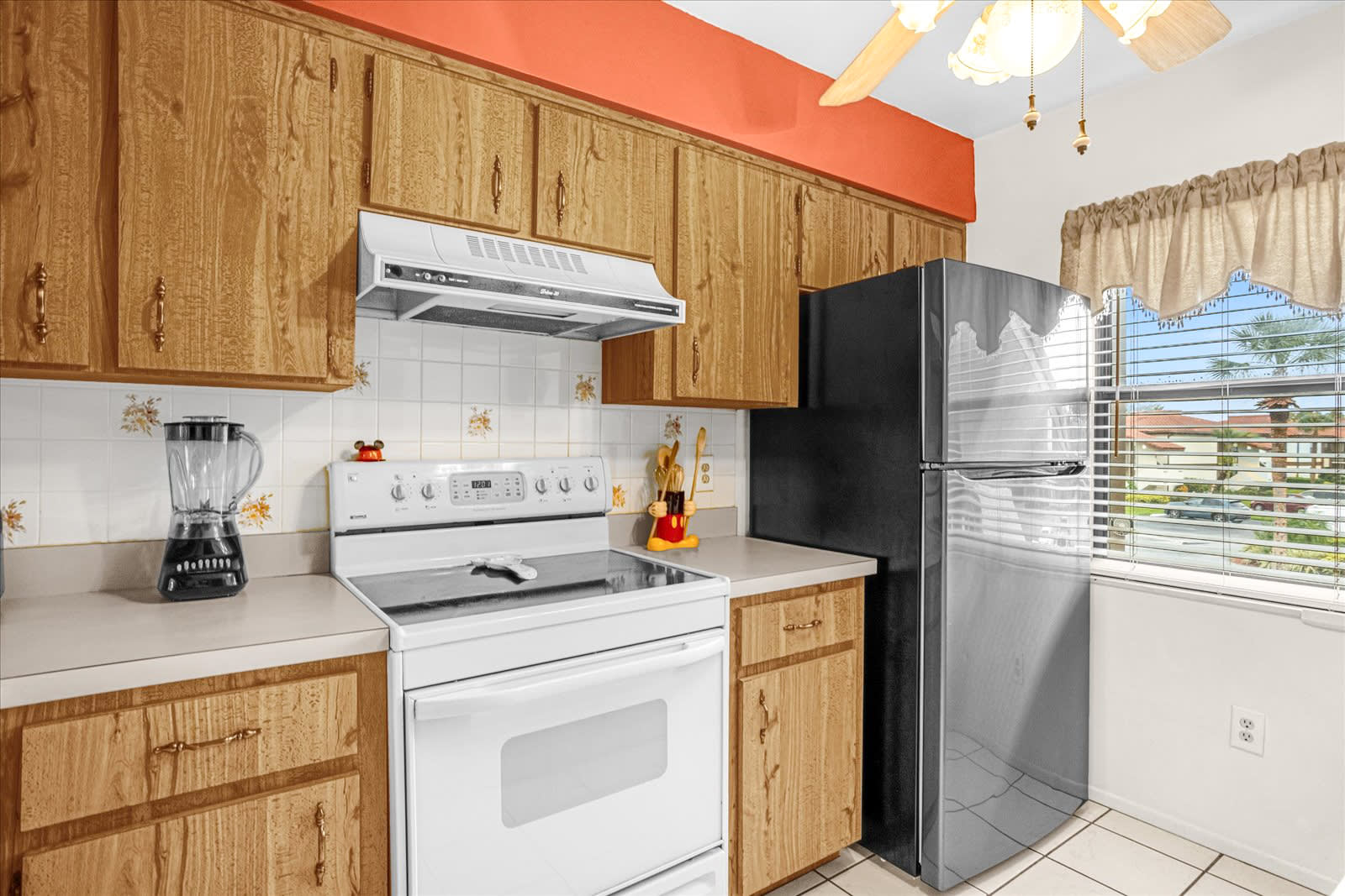 This kitchen showcases light wood cabinets, a white tile backsplash with subtle floral patterns, and a vibrant orange accent wall. It features a white electric stove with an overhead range hood, a black and stainless steel refrigerator, and a window with blinds.