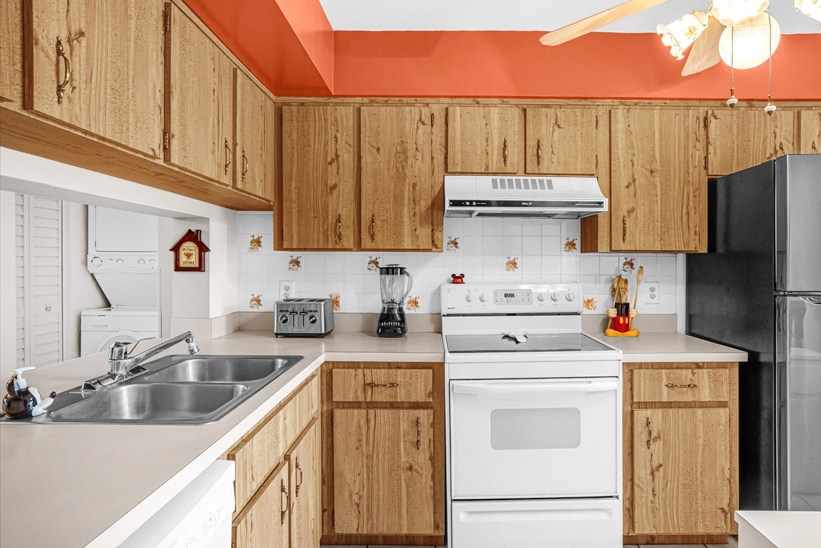 A bright kitchen features light wood cabinets, white tile backsplash with small floral accents, and an orange accent wall. It includes a double sink, white appliances, a black refrigerator, and a laundry area visible through an opening.