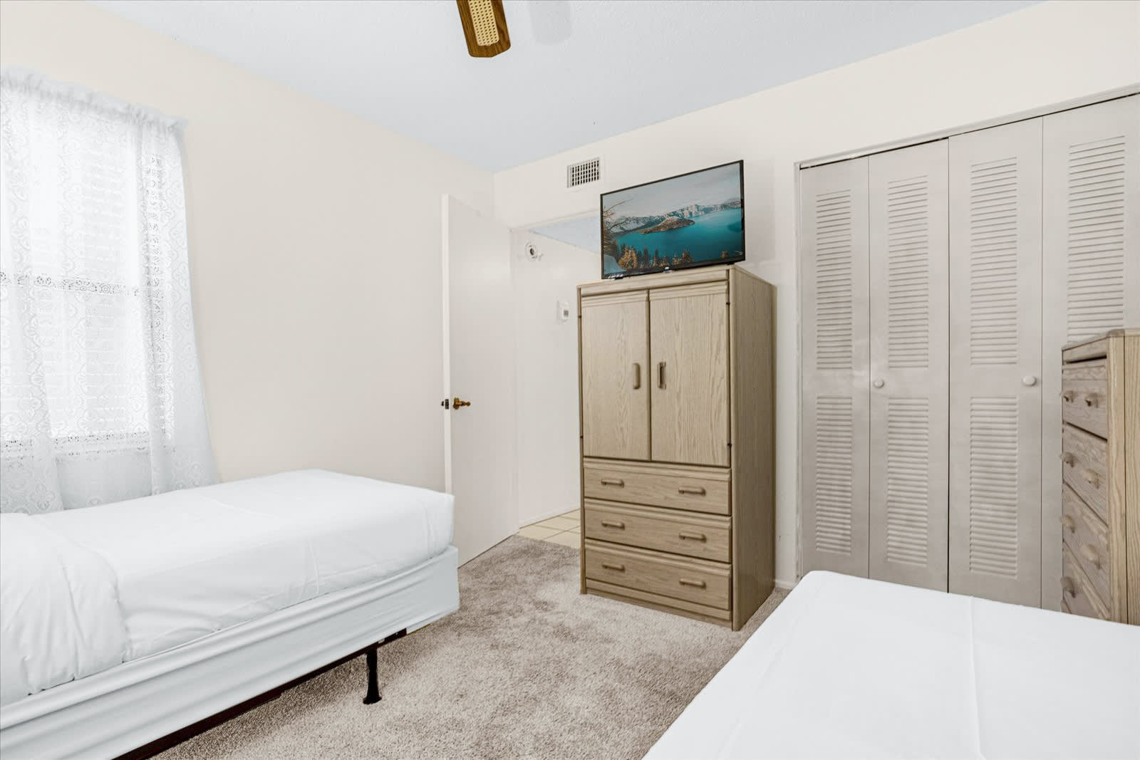 A bedroom with two white twin beds, light beige carpet, and a light-colored armoire with a flat-screen TV mounted above it. A closet with bi-fold doors is visible, and a window with sheer curtains brightens the space.