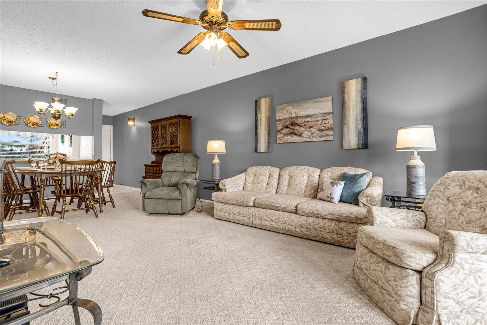 An open-concept living and dining area with a patterned beige sofa and armchair, light beige carpet, and gray walls, featuring a wooden hutch and a round dining table in the background.