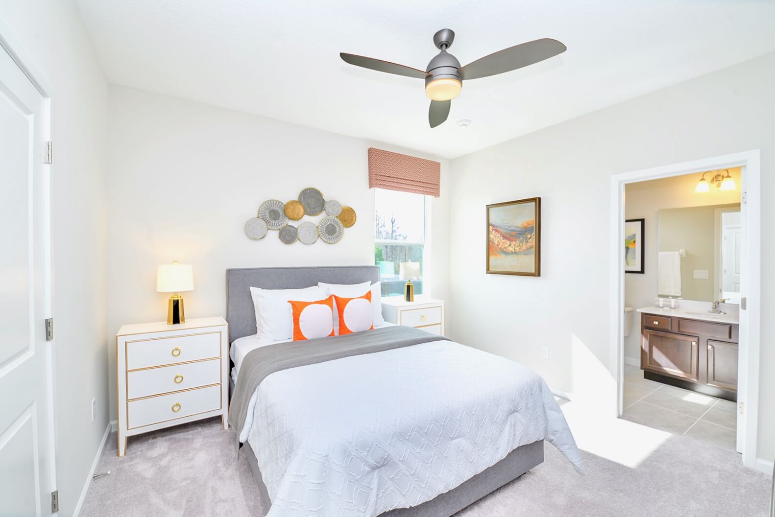 Modern bedroom featuring a queen-sized bed with a gray upholstered headboard, white bedding, and two orange accent pillows. Two white nightstands with gold hardware flank the bed, and a ceiling fan hangs overhead.