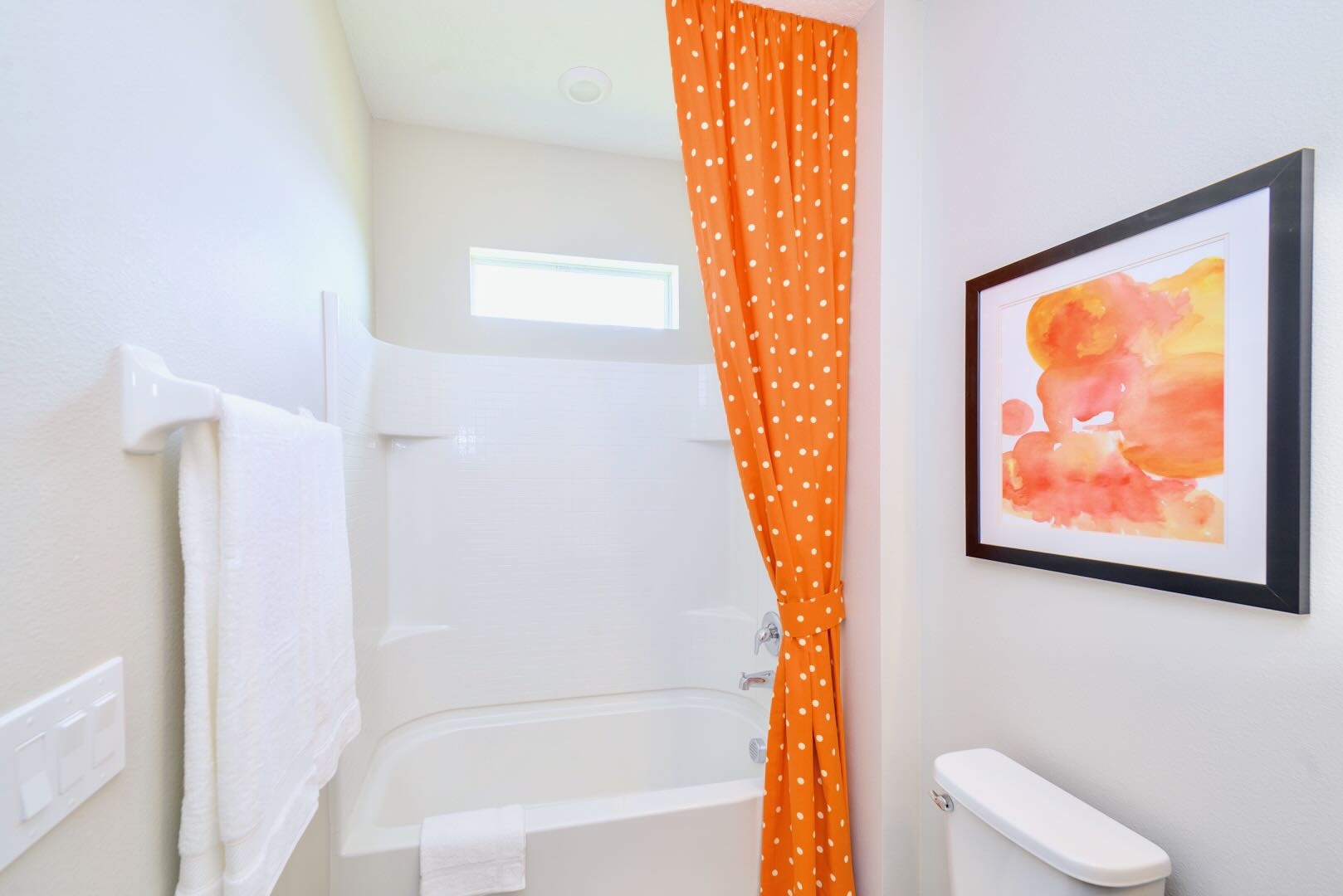 Small bathroom with a white toilet and a white bathtub/shower combo. An orange polka-dot shower curtain and a framed abstract art piece with orange and yellow hues add a playful touch.