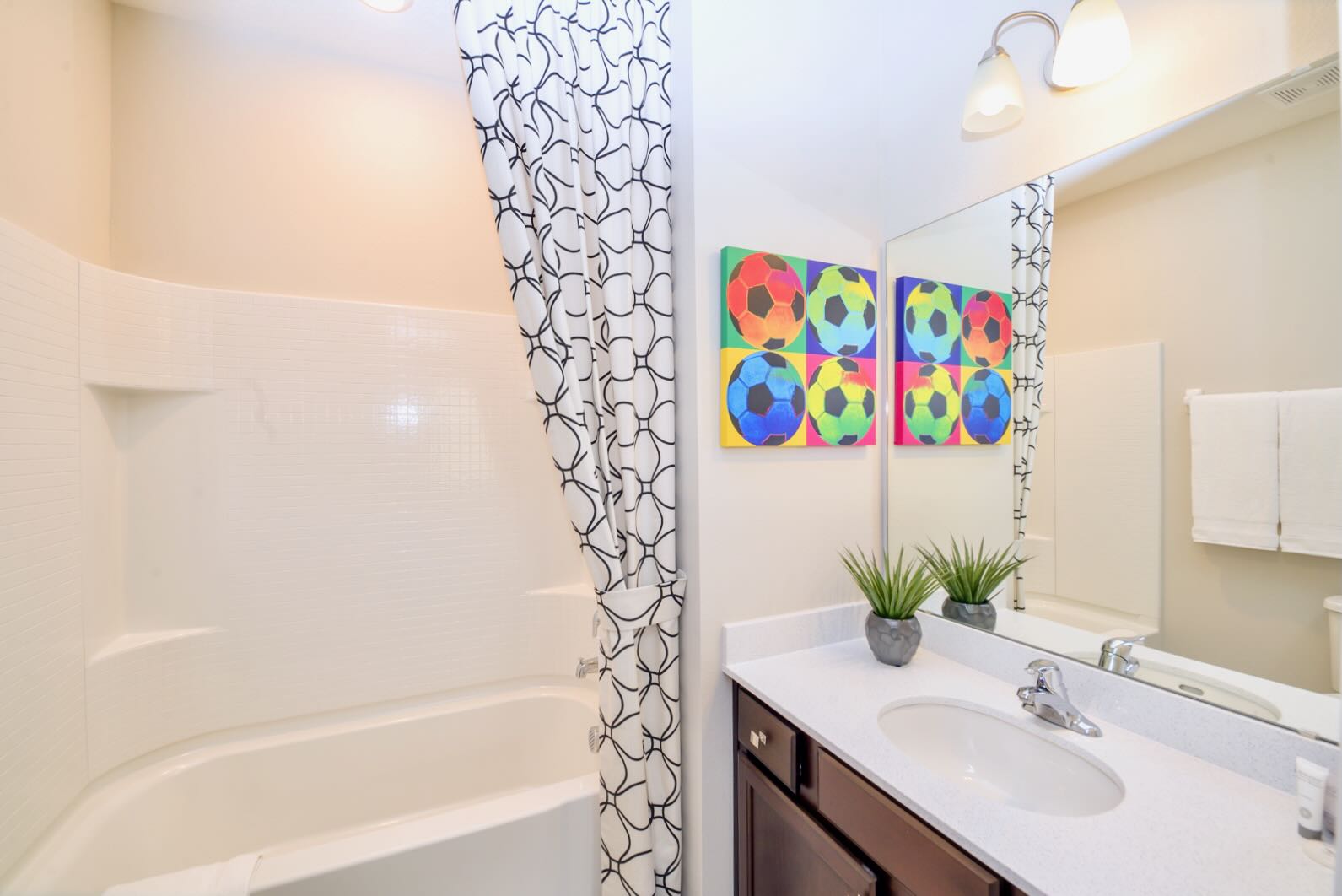 Modern bathroom with a dark wood vanity, single sink, large mirror, and two-light fixture. A colorful pop-art style soccer ball print hangs above the toilet, complementing the black and white patterned shower curtain.