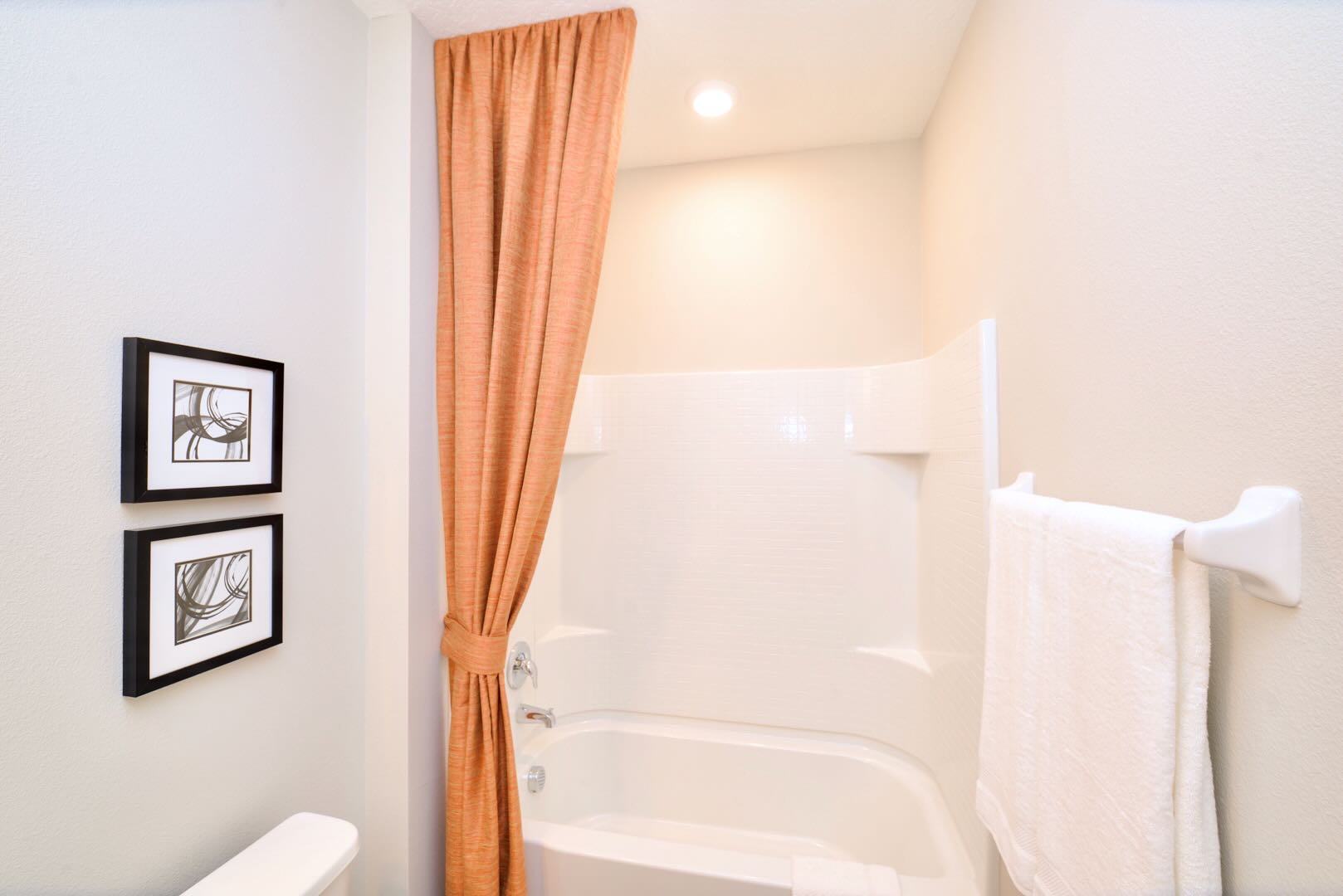 Simple bathroom featuring a white shower/tub combo with an orange shower curtain tied back, and a toilet. Two framed abstract artworks are mounted on the wall above the toilet.