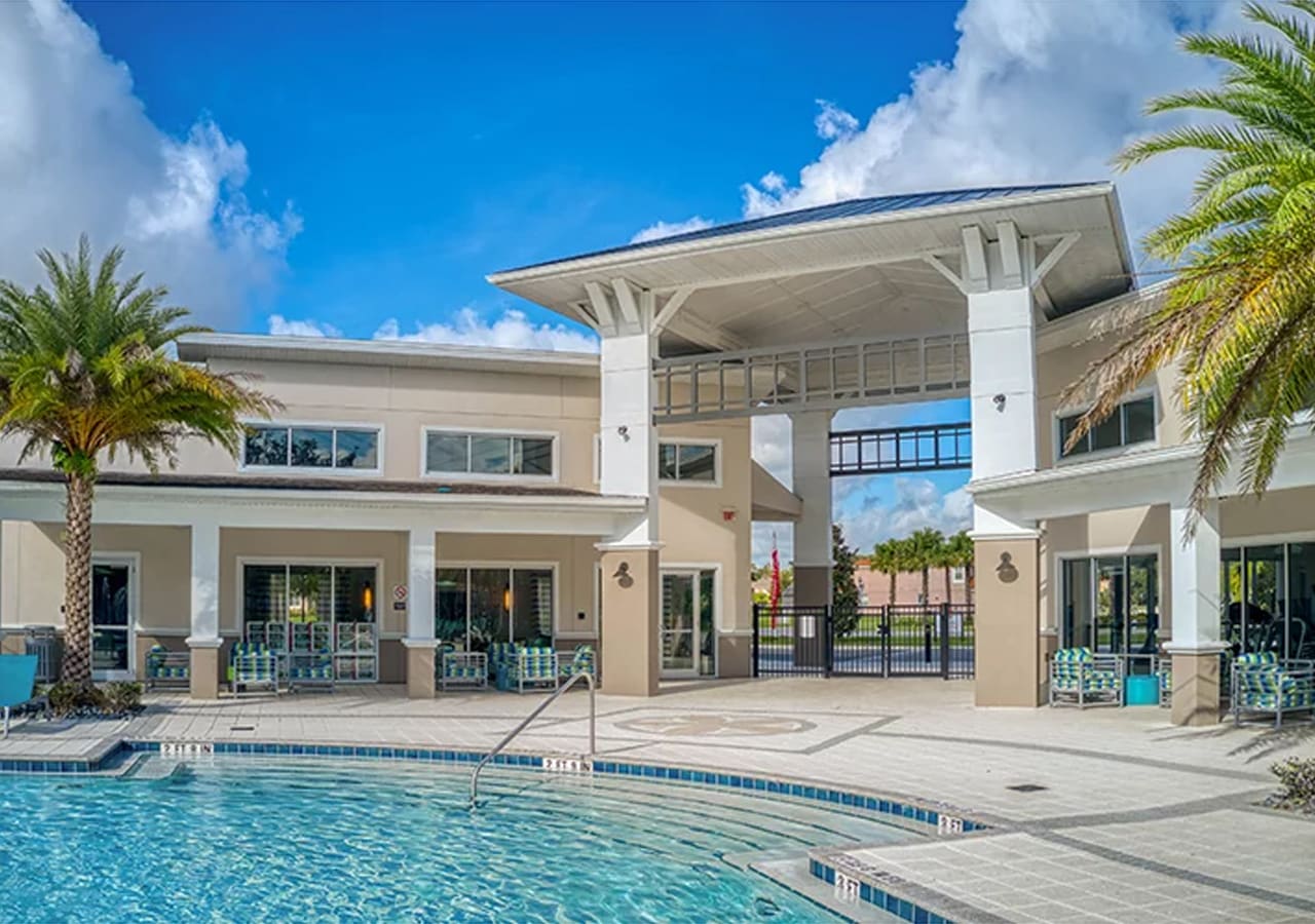 Expansive communal pool area with a large swimming pool, surrounded by a paved deck and lounge chairs. A grand clubhouse building with columns and palm trees is visible in the background under a bright blue sky.