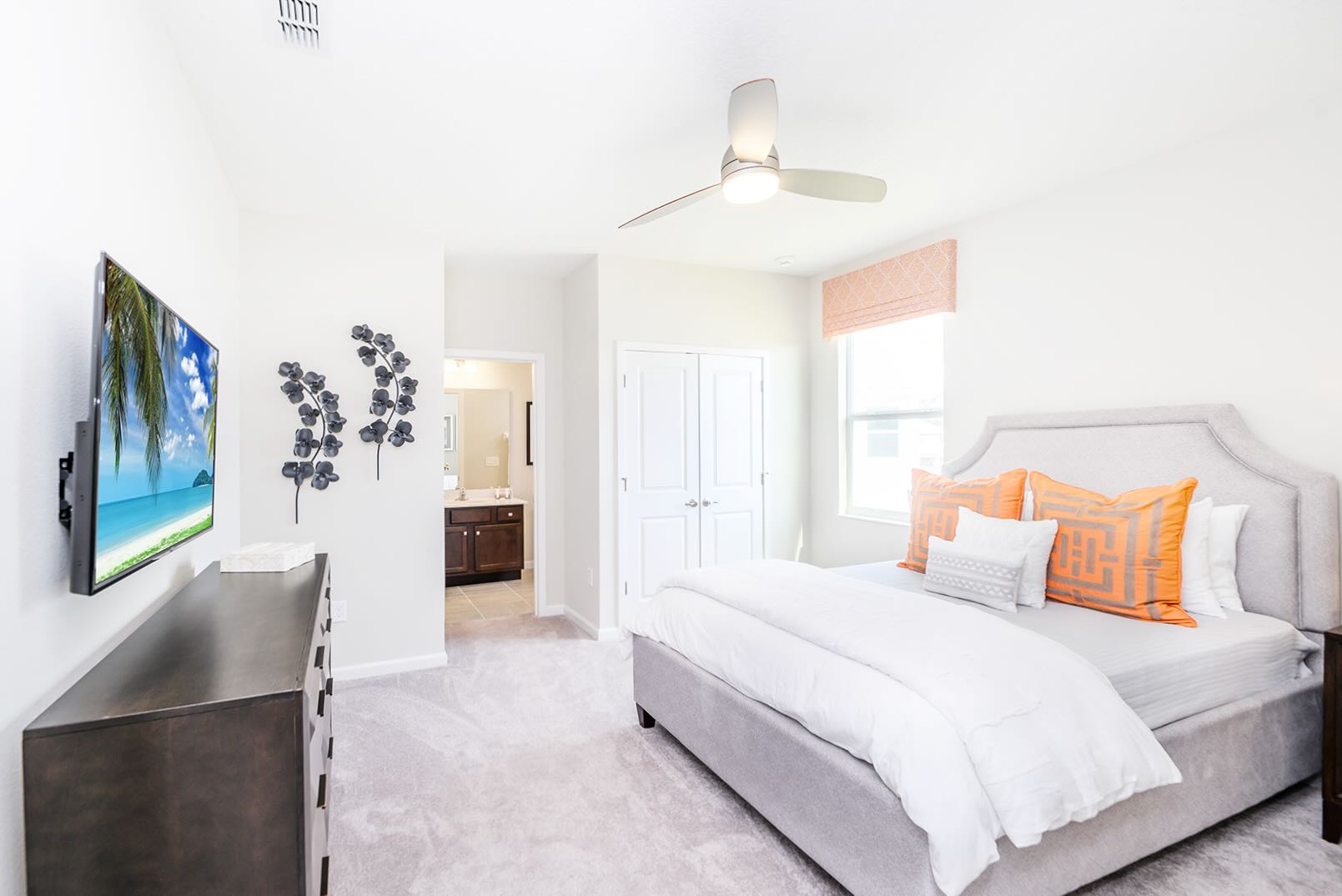 Spacious bedroom with a gray upholstered bed, white bedding, and orange accent pillows. A wall-mounted TV, a dark wood dresser, and a view into an en-suite bathroom are visible, offering comfort and convenience.
