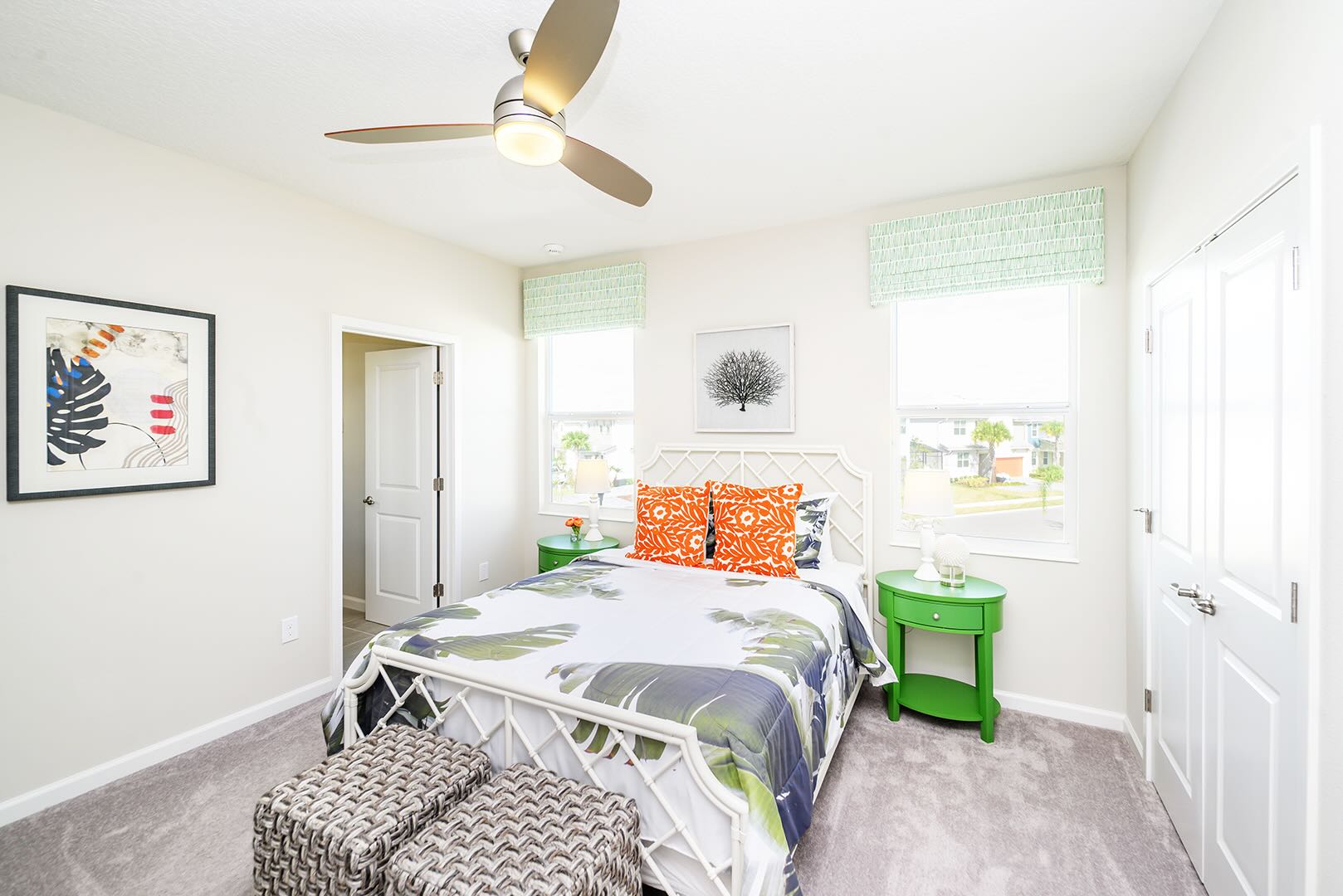 Bright bedroom with a white metal bed frame, tropical leaf patterned bedding, and orange accent pillows. Two green round nightstands with lamps, a ceiling fan, and two woven ottomans are present, creating a vibrant and inviting space.