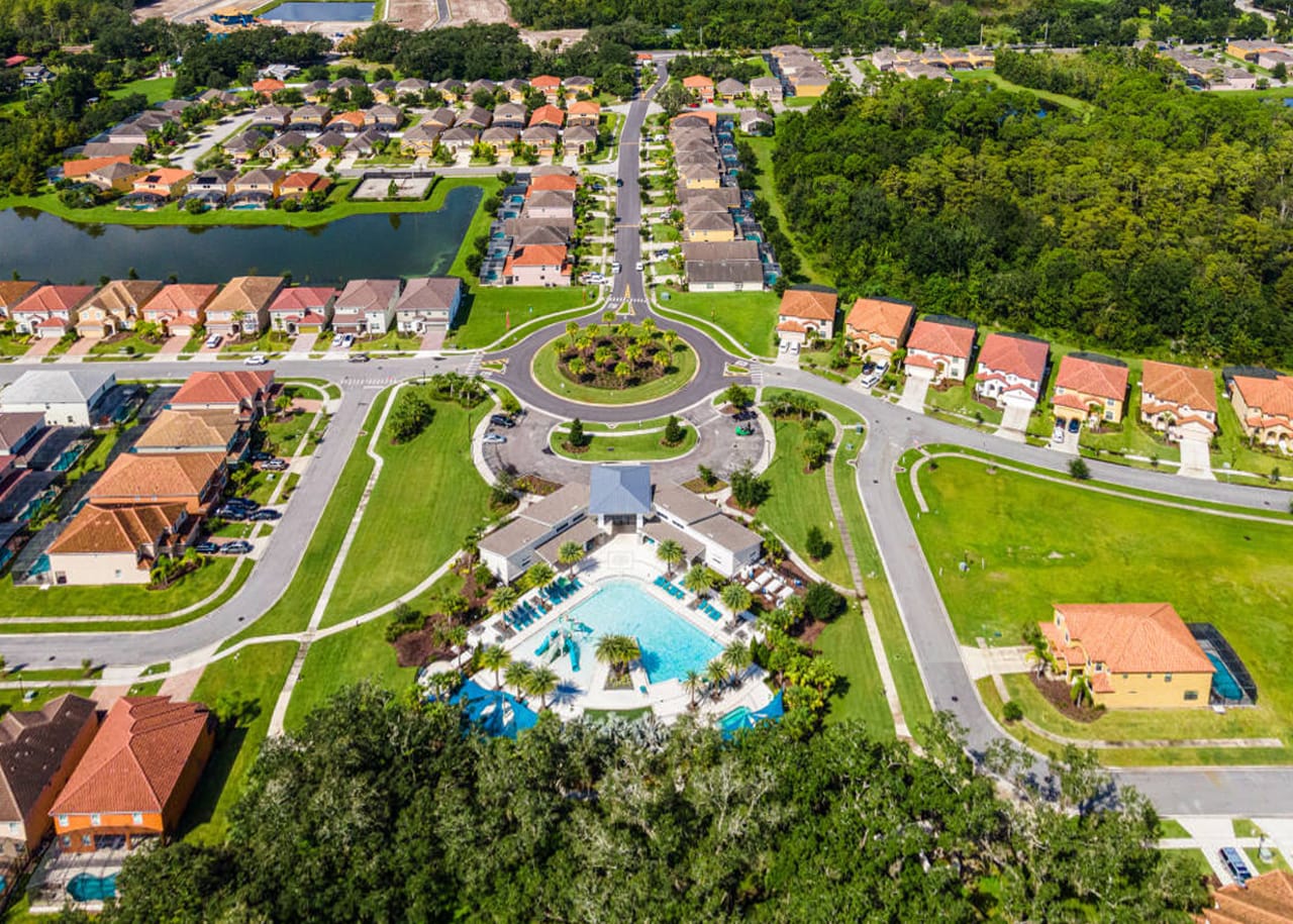 High aerial view of a residential community featuring numerous houses with terracotta roofs, winding roads, a central clubhouse with a large communal pool area, and surrounding green spaces and water bodies.