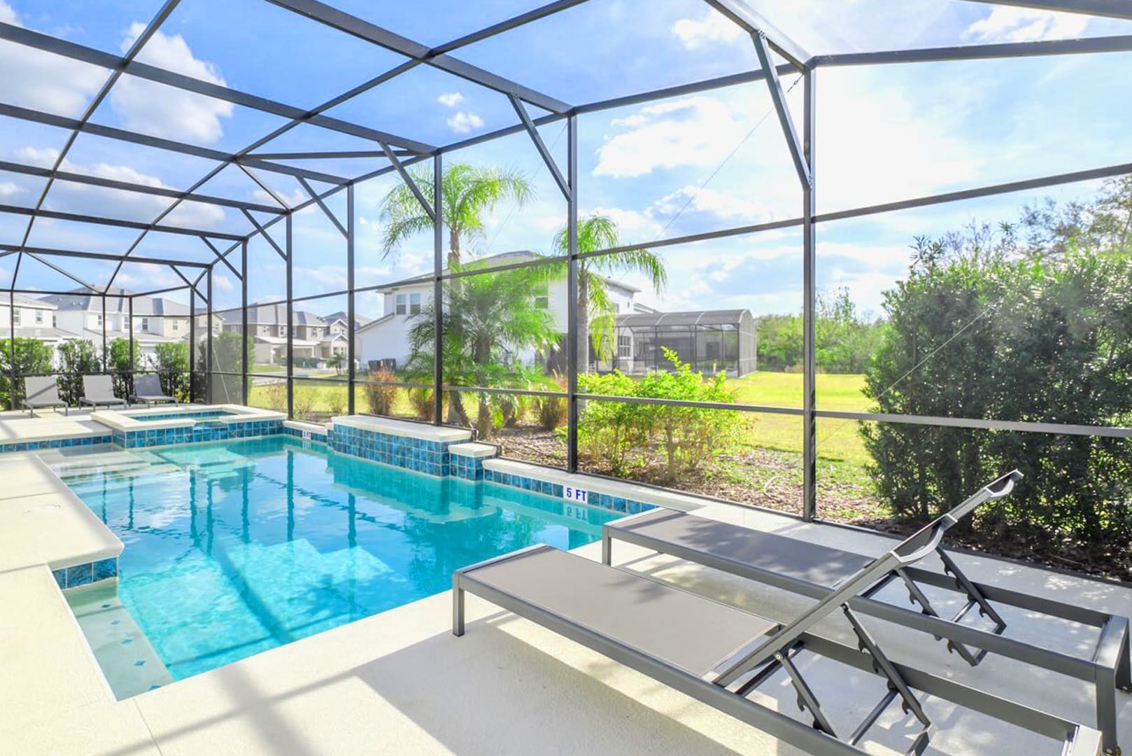 A screened-in private pool area with a rectangular swimming pool and an integrated hot tub, surrounded by light-colored decking and two lounge chairs. The enclosure offers views of a grassy backyard and neighboring houses.