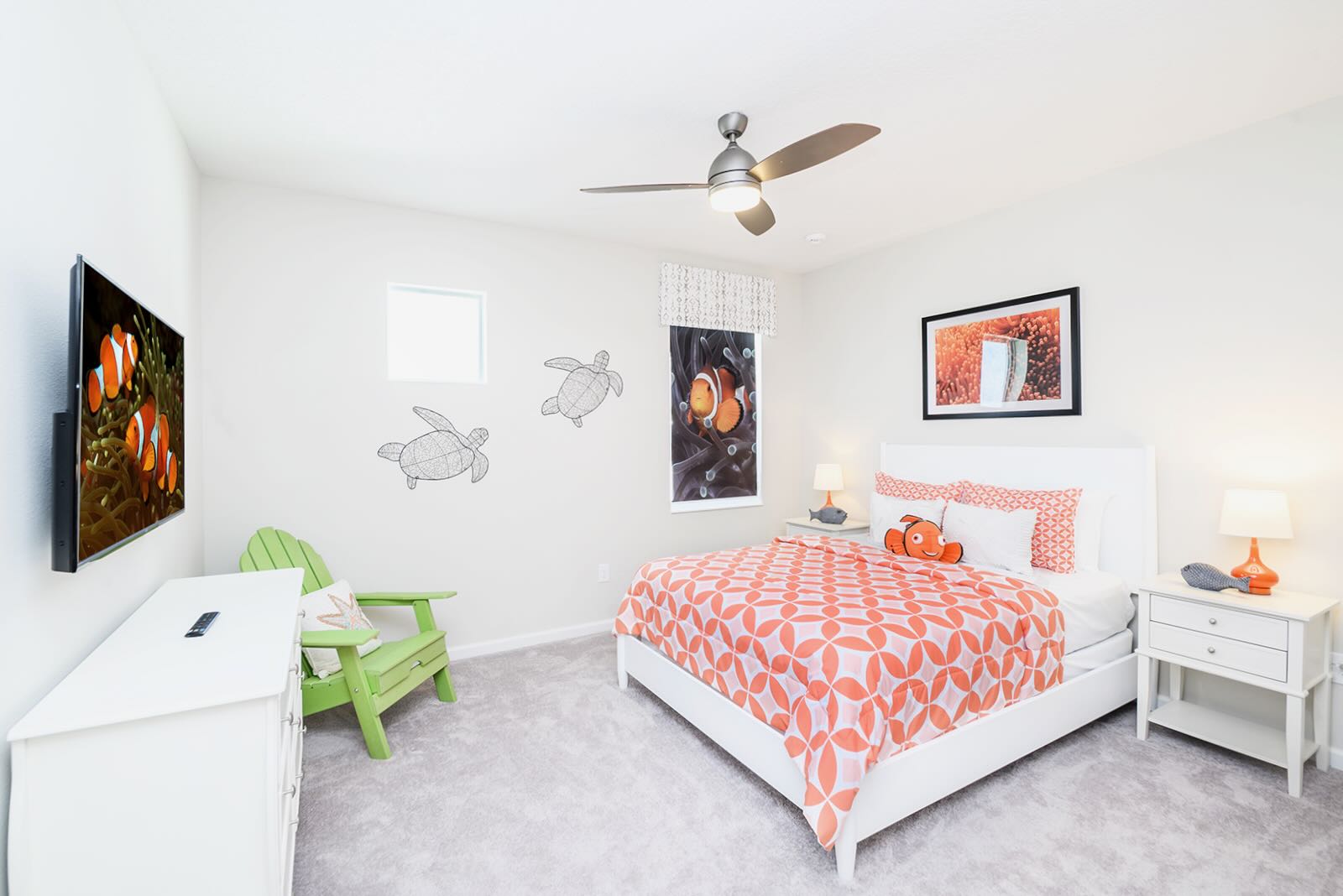 Kids' bedroom with a white bed featuring orange patterned bedding and a Nemo-themed pillow. A wall-mounted TV, a green Adirondack chair, and clownfish-themed decor are present, creating a playful atmosphere.