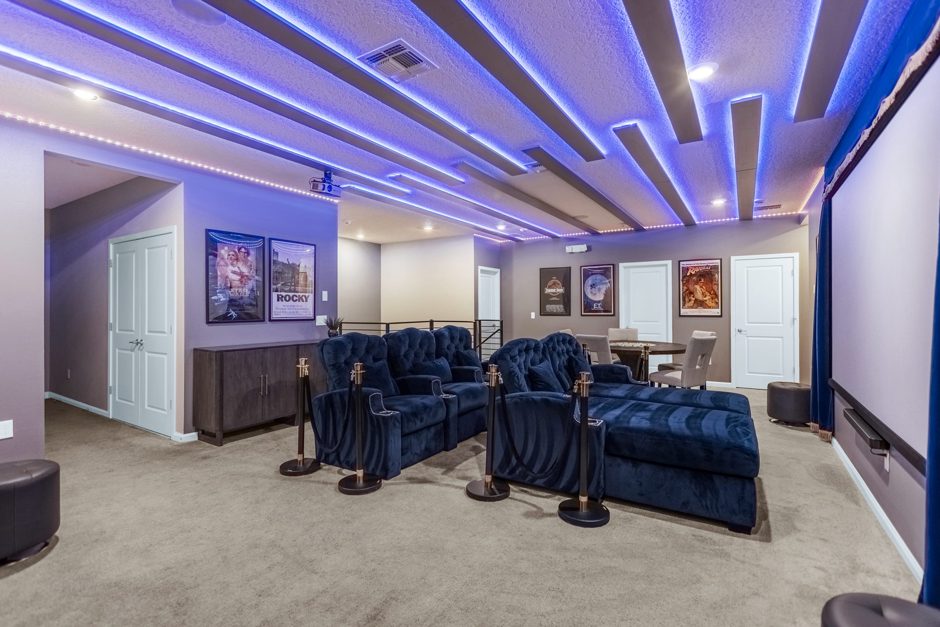 Spacious media room featuring multiple rows of plush blue reclining theater seats, a large projection screen, and a round game table. Blue LED strip lighting accents the modern ceiling design, with movie posters adorning the walls.