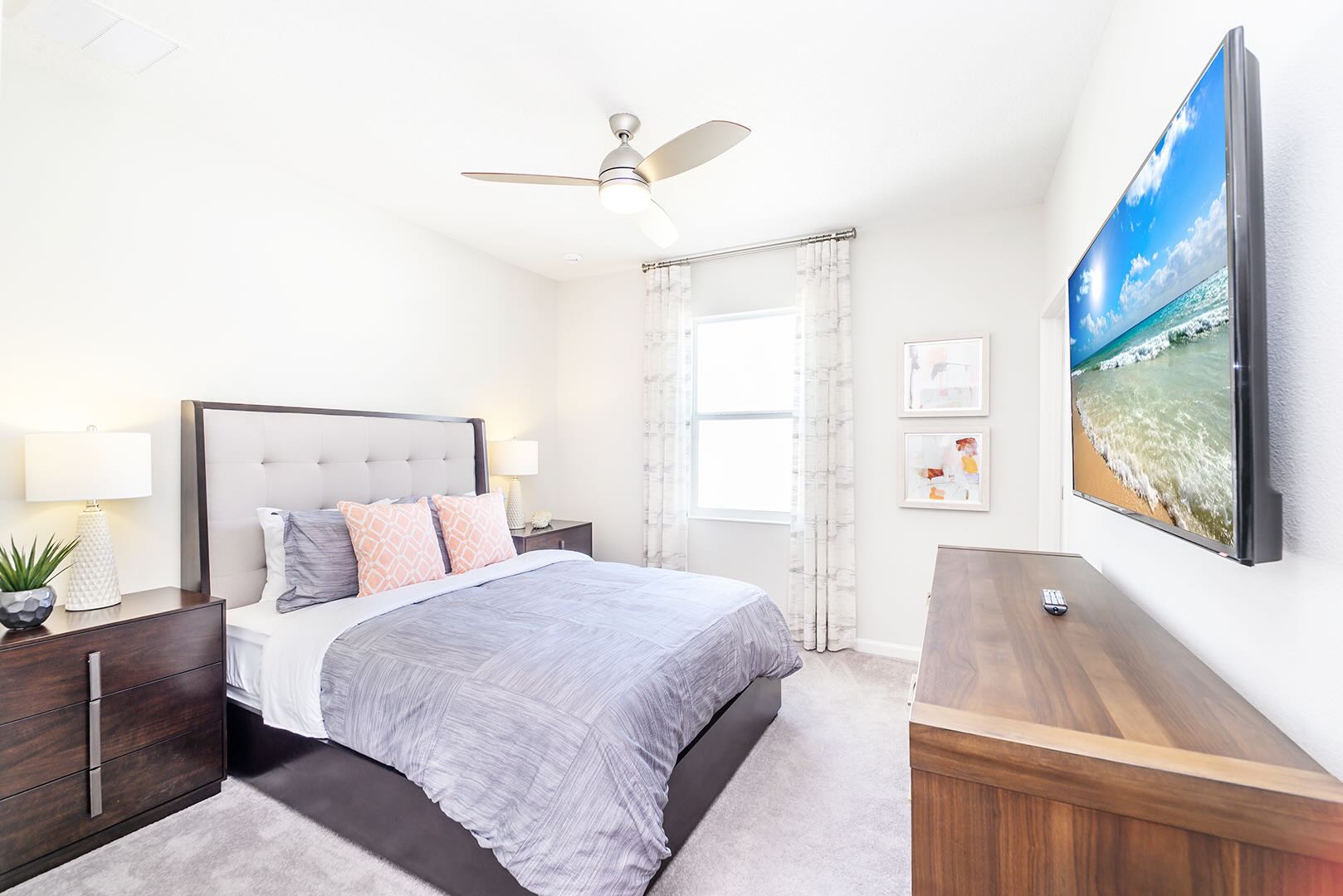 Bright bedroom with a queen-sized bed featuring a gray upholstered headboard and gray bedding with coral accent pillows. A ceiling fan, two dark wood nightstands with lamps, and a wall-mounted TV are visible.