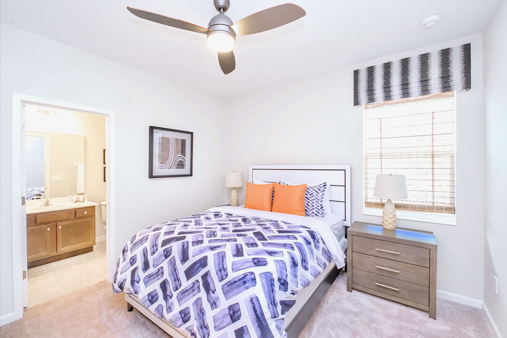 Cozy bedroom with a queen bed featuring a white and gray headboard, a blue and white patterned duvet, and orange accent pillows. A light wood nightstand with a lamp sits beside the bed, and a ceiling fan is overhead.