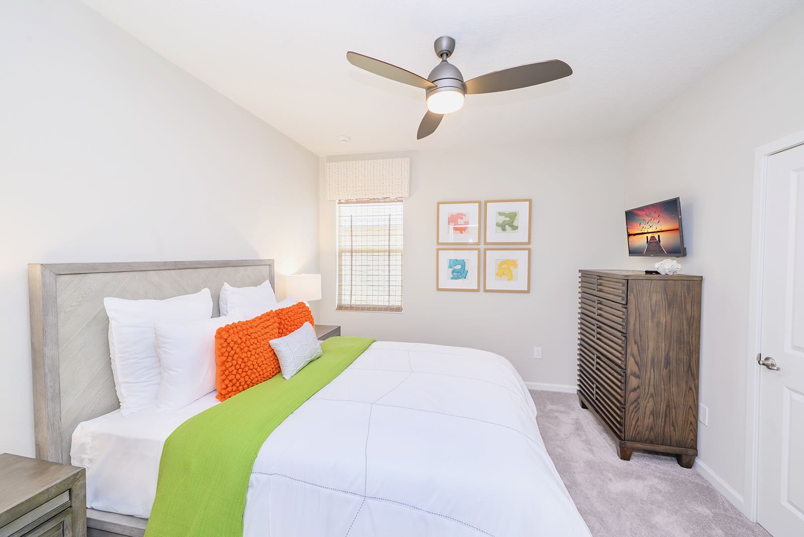 Bright bedroom with a queen bed featuring a light wood headboard, white bedding, and a vibrant green throw blanket with orange accent pillows. A wall-mounted TV is opposite the bed, and a ceiling fan provides air circulation.