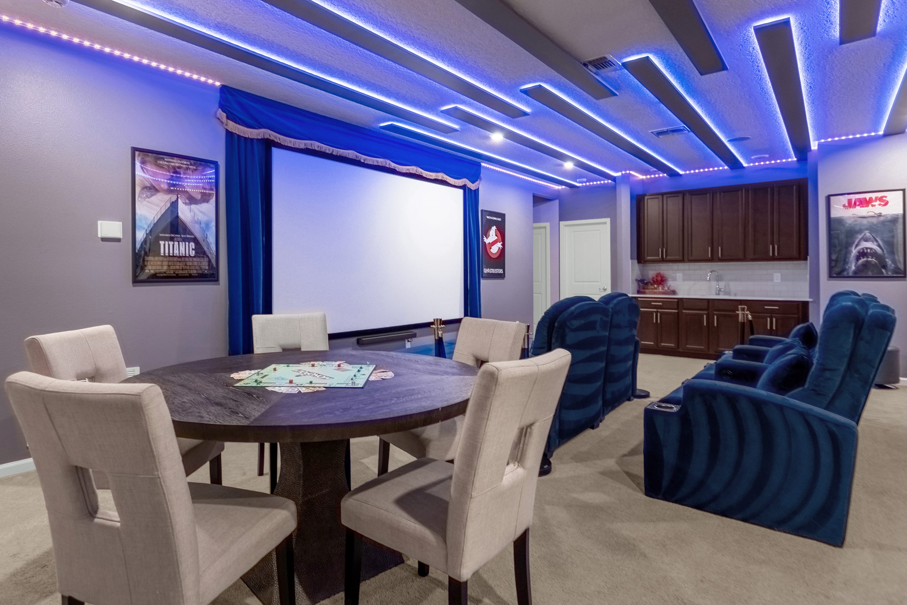 Cozy media room with a large projection screen, plush blue reclining theater seats, and a round table with chairs for board games. A dark wood wet bar with upper cabinets is visible in the background, all under blue LED ceiling lights.