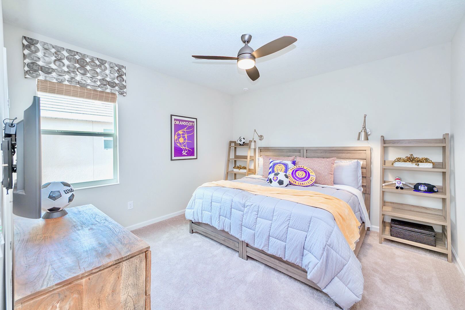 Soccer-themed bedroom with a queen-sized bed, gray bedding, and purple and yellow accent pillows. A wall-mounted TV, a wooden dresser, and two tall wooden shelving units flank the bed, creating a sporty and organized space.