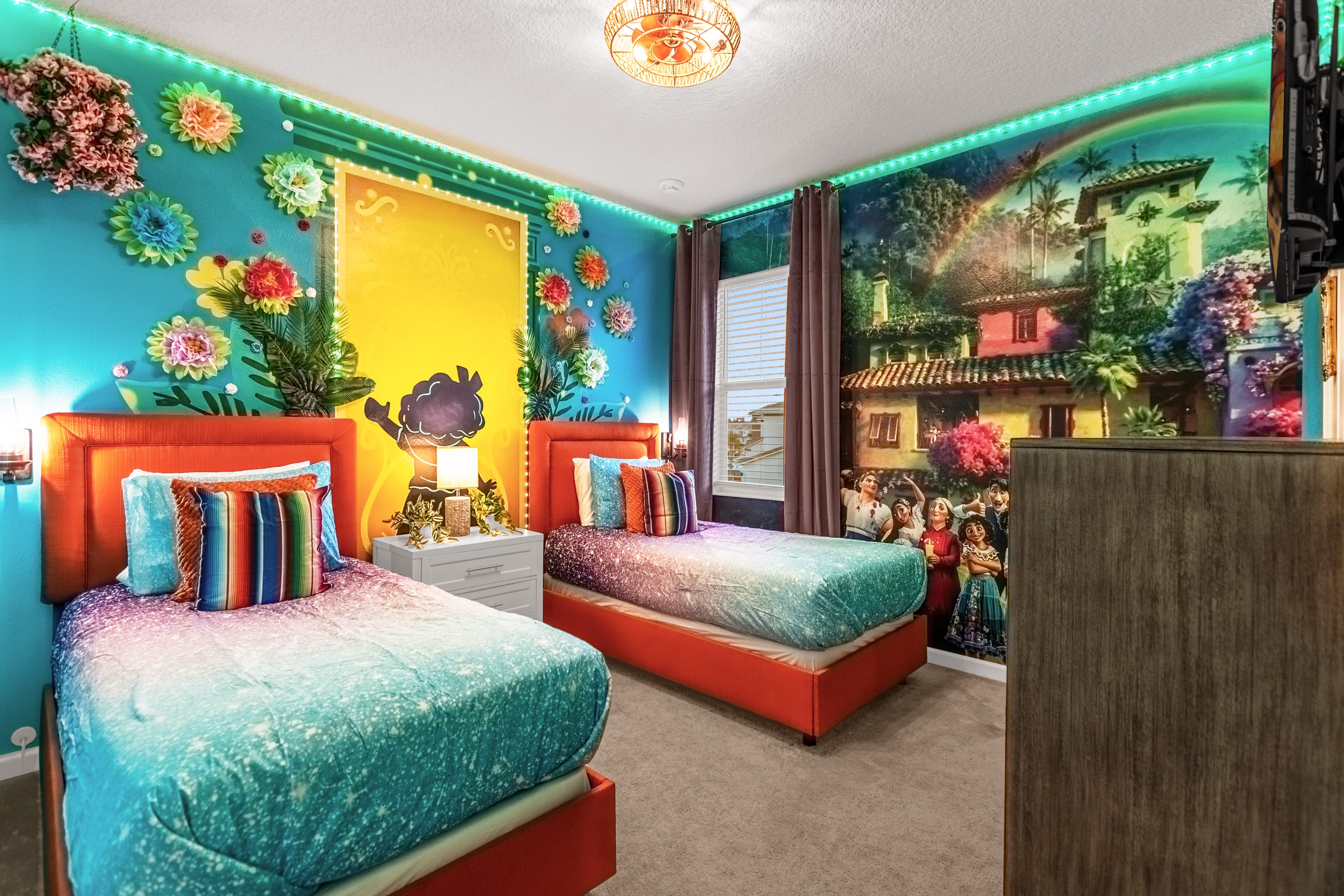 Vibrant themed bedroom with two twin beds featuring orange frames and colorful bedding, set against a bright blue wall with floral and mural decorations, and green LED strip lighting.