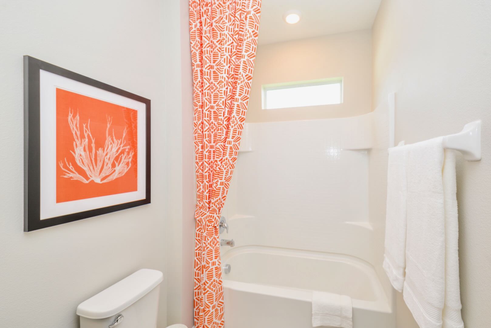 Bright bathroom with a white toilet and a white bathtub/shower combo. An orange and white patterned shower curtain and a framed coral art piece provide a pop of color and a coastal feel.