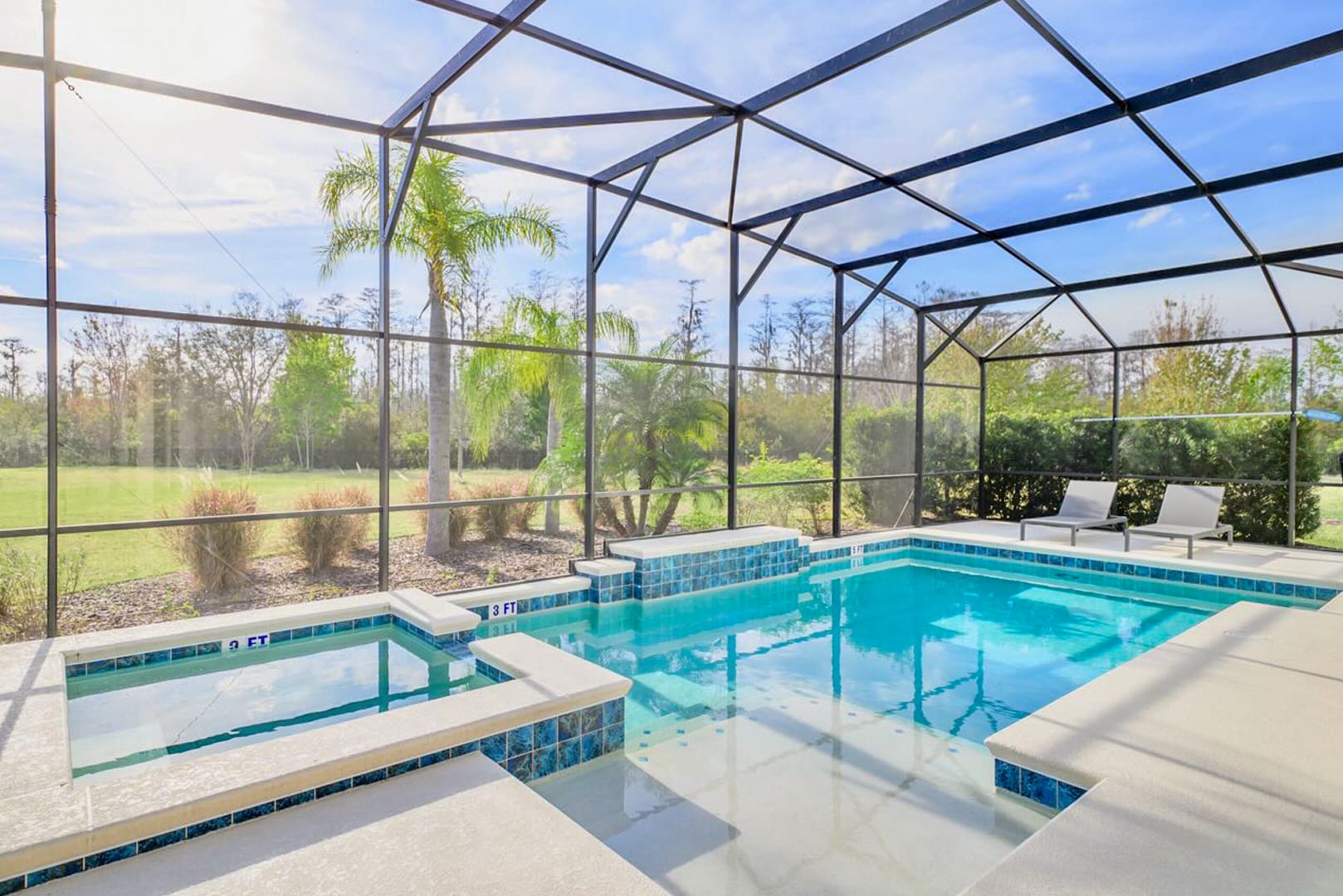 A screened-in outdoor pool area features a rectangular swimming pool with an integrated hot tub, surrounded by light-colored decking. Palm trees and lush greenery are visible beyond the screen, under a bright blue sky.