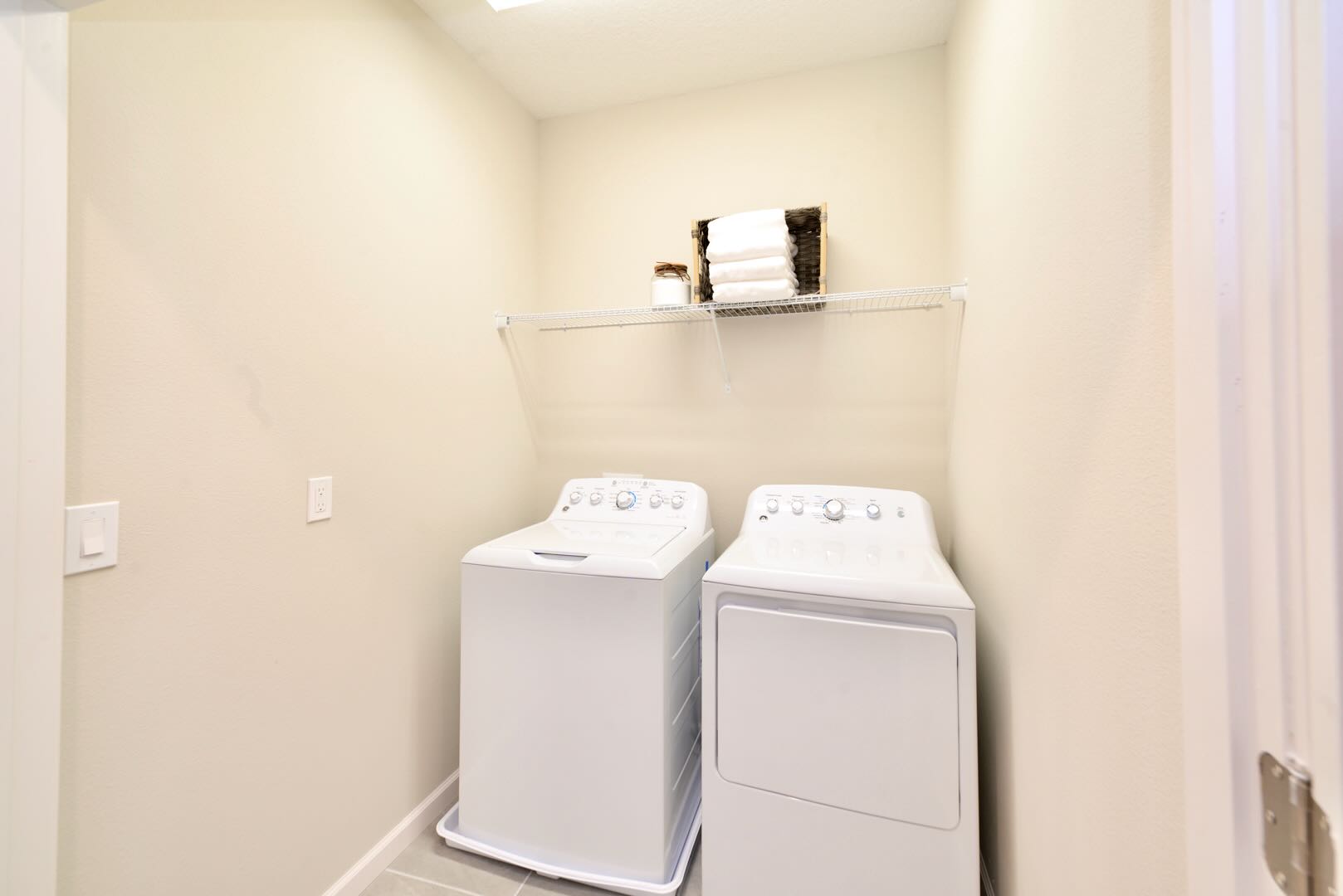 Functional laundry room equipped with a white top-loading washing machine and a matching dryer. A wire shelf above provides storage space for towels and laundry essentials.