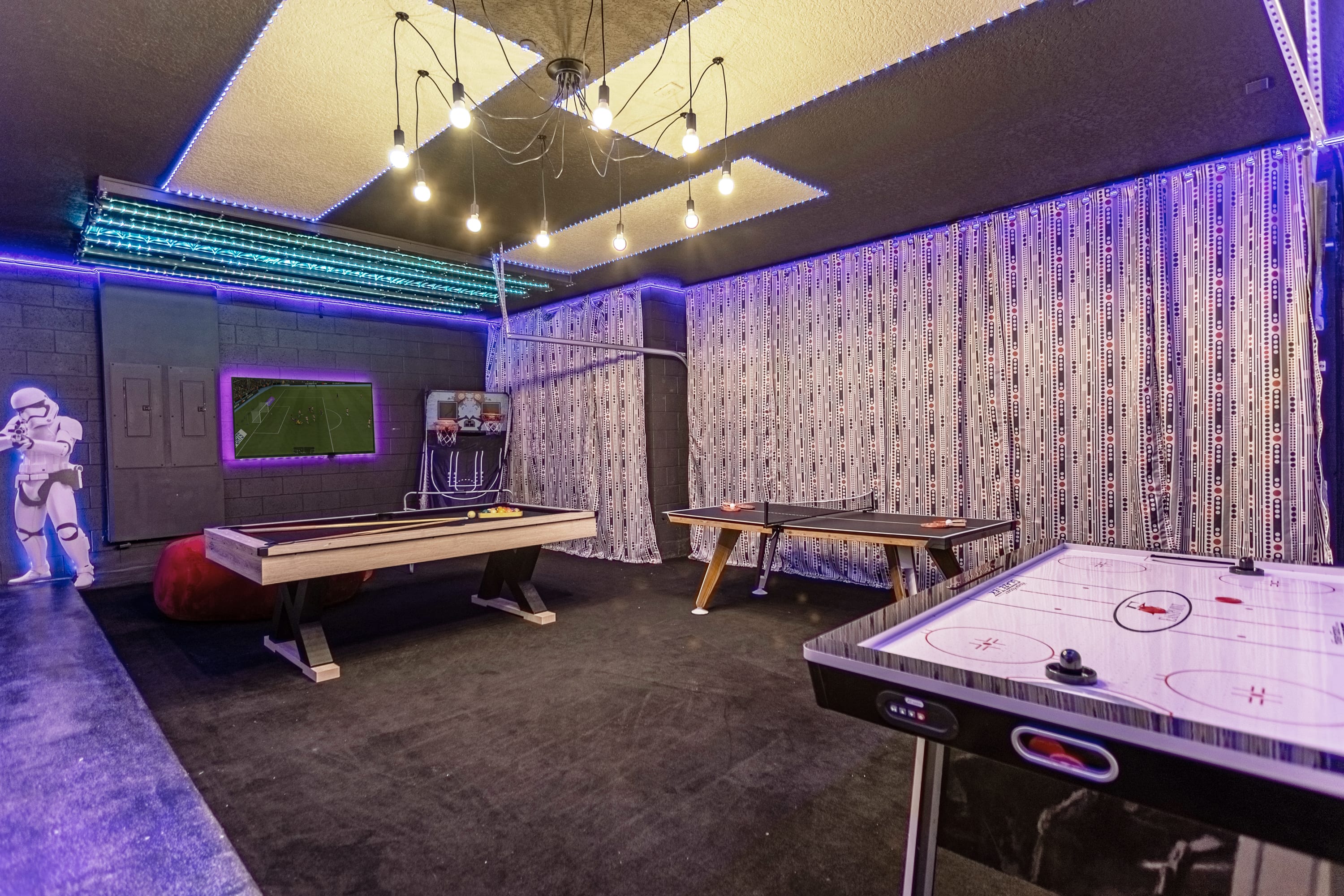 Vibrant game room featuring a pool table, air hockey table, and basketball arcade game, illuminated by blue LED strip lighting and a unique ceiling light fixture. A large stormtrooper statue stands guard in the corner.