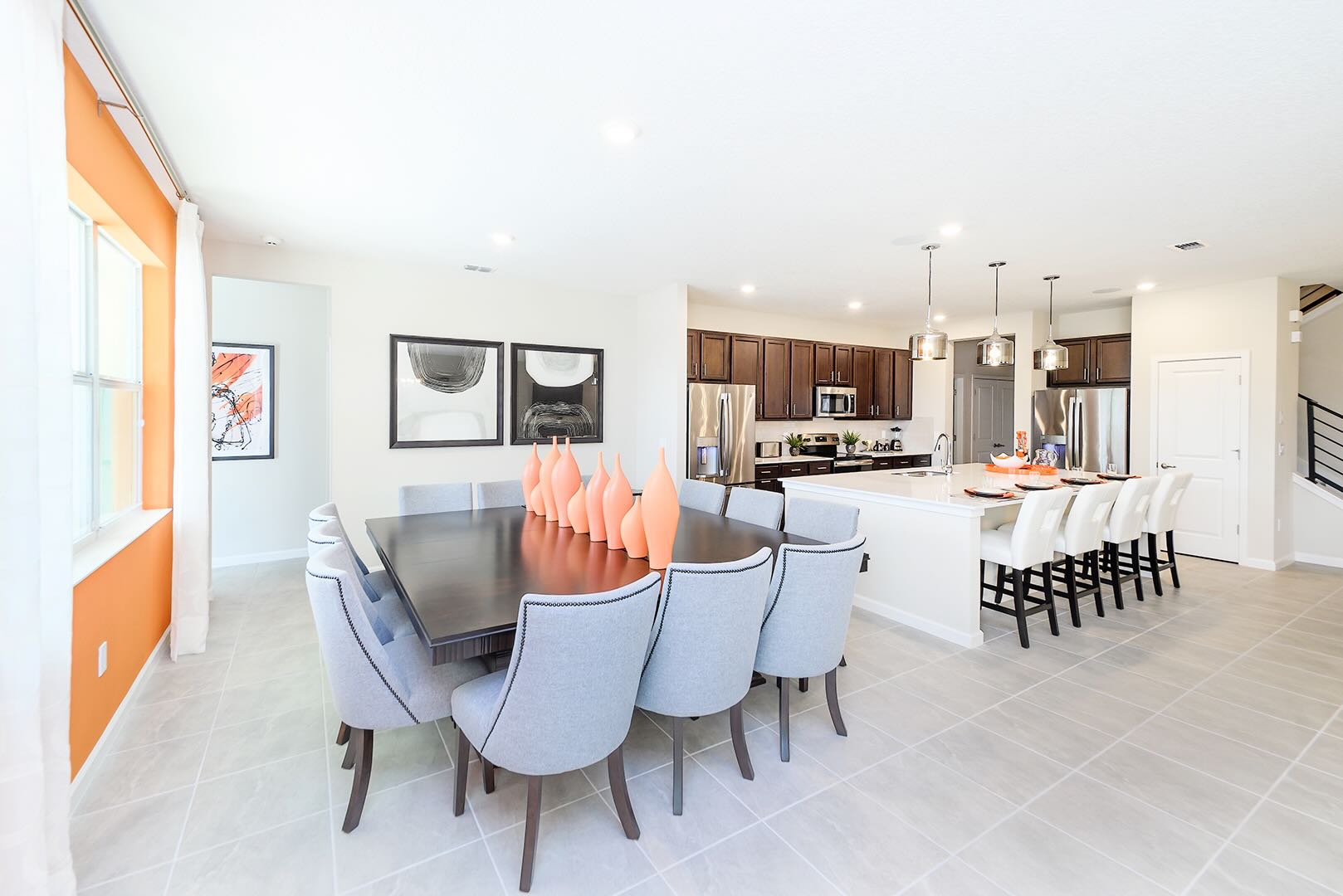 A contemporary dining room with a large dark wood table surrounded by ten upholstered chairs, decorated with orange vases. It's part of an open-concept space, flowing into a modern kitchen with dark wood cabinets and a breakfast bar.