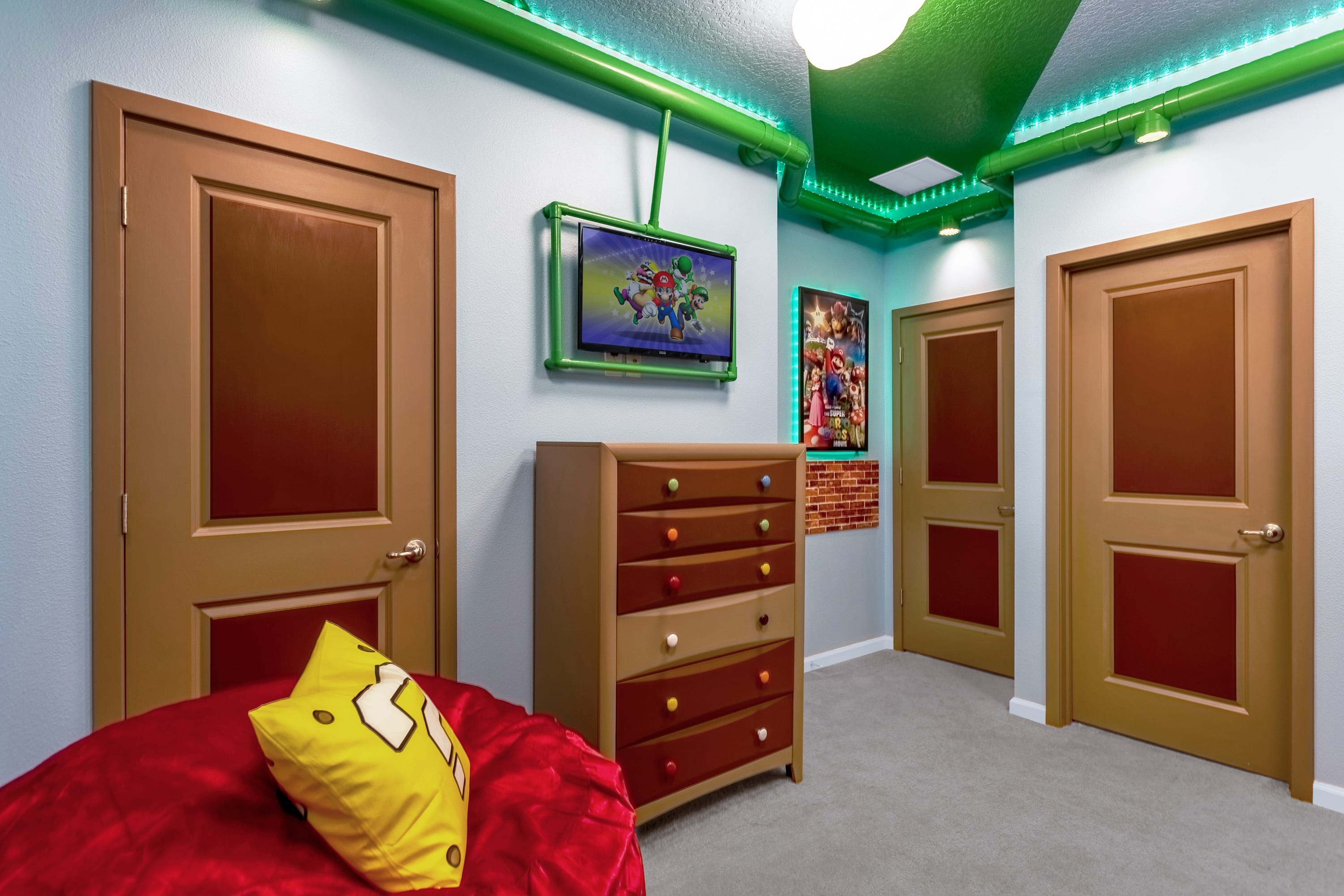 Themed bedroom with a colorful dresser and a wall-mounted TV, featuring green pipe-like ceiling decor with LED lighting and multiple brown doors with gold panels.