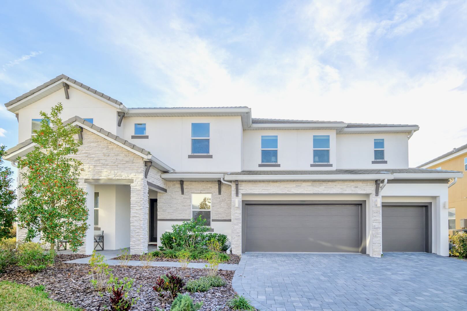 A modern two-story house with a white and light stone facade, featuring multiple windows and a two-car garage with a gray door. The front yard has low-maintenance landscaping with small trees and shrubs, and a paved driveway.