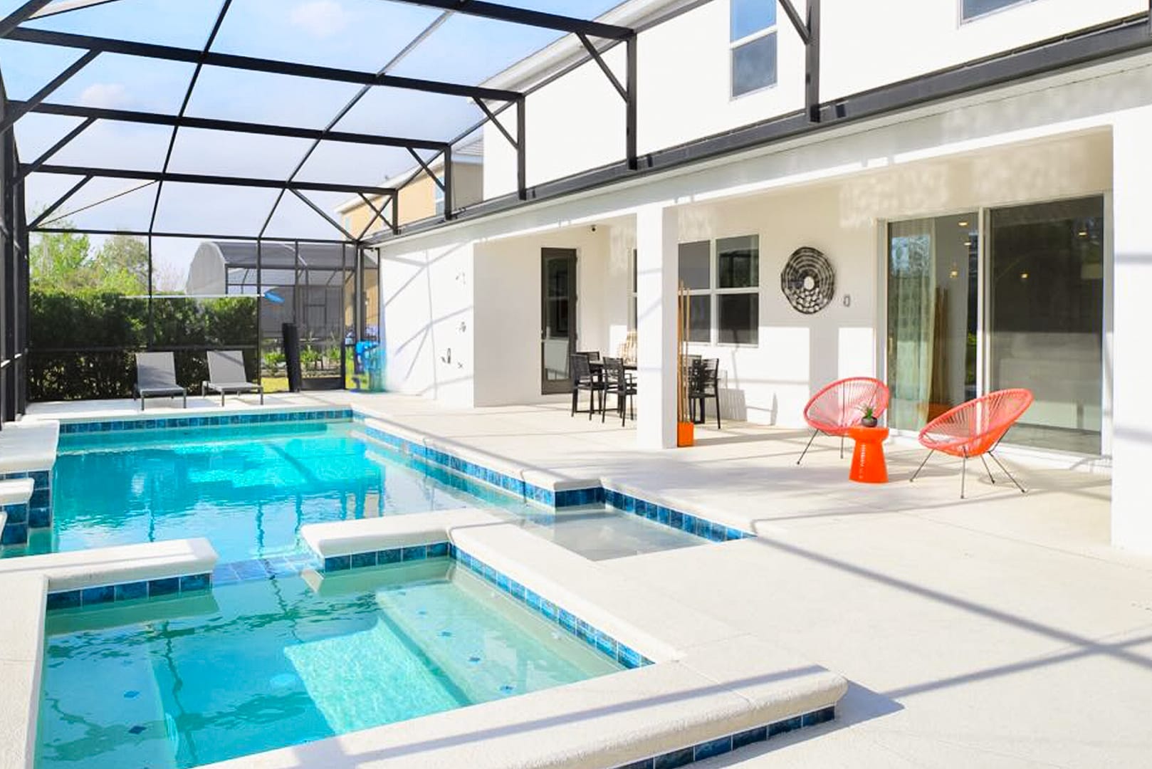A wide view of the screened-in pool area, showing the rectangular pool and hot tub, with a covered patio area adjacent to the house. The patio features two modern orange chairs and a small table, with views of the surrounding landscape.