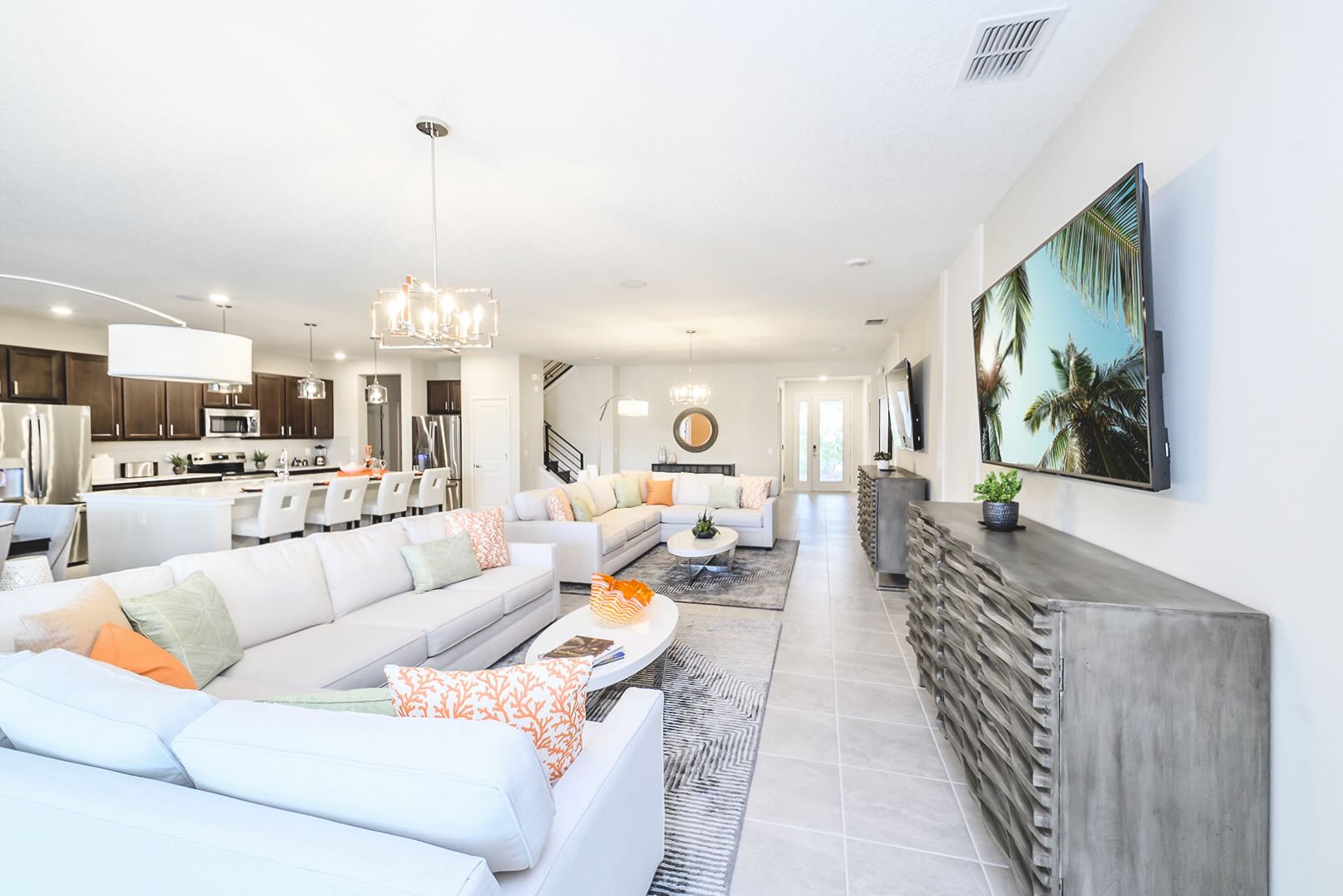 Bright and expansive open-concept living room with a large white sectional sofa, a modern coffee table, and a wall-mounted TV above a textured console. Light tile flooring extends throughout the space, leading to the kitchen in the background.