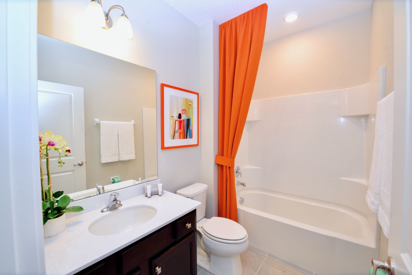 Modern bathroom with a dark wood vanity, single sink, white countertop, and a shower/tub combo with a bright orange shower curtain. A framed abstract art piece adds a pop of color to the wall.