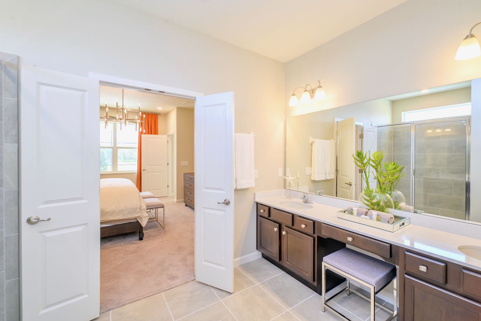 Modern master bathroom featuring a long double vanity with dark wood cabinets, light countertops, and a large mirror, with a view into a carpeted bedroom through an open doorway.