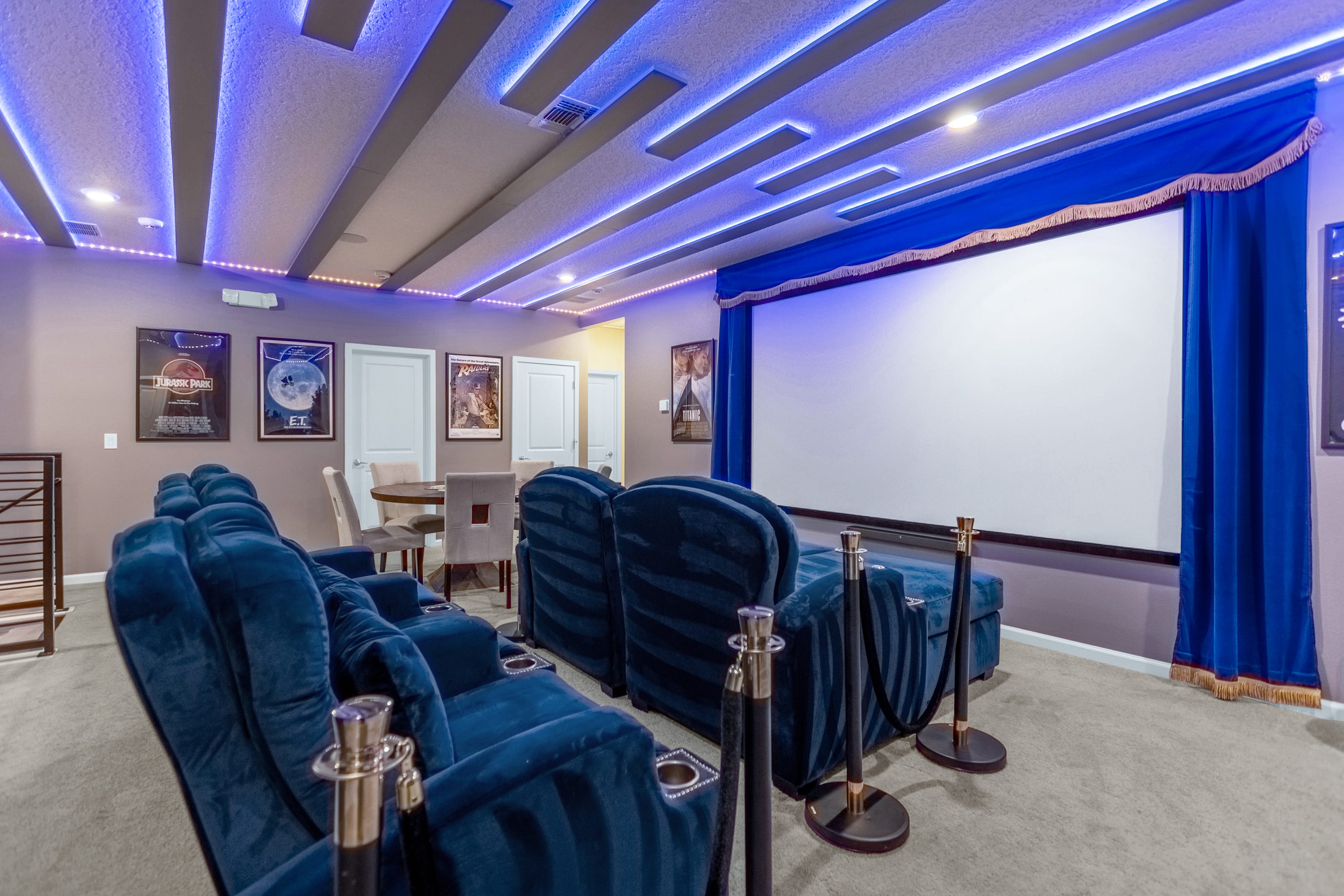 A dedicated home theater features rows of plush blue velvet recliners with cup holders, facing a large projection screen framed by blue curtains. The ceiling is adorned with linear blue LED strip lighting, creating a cinematic ambiance.