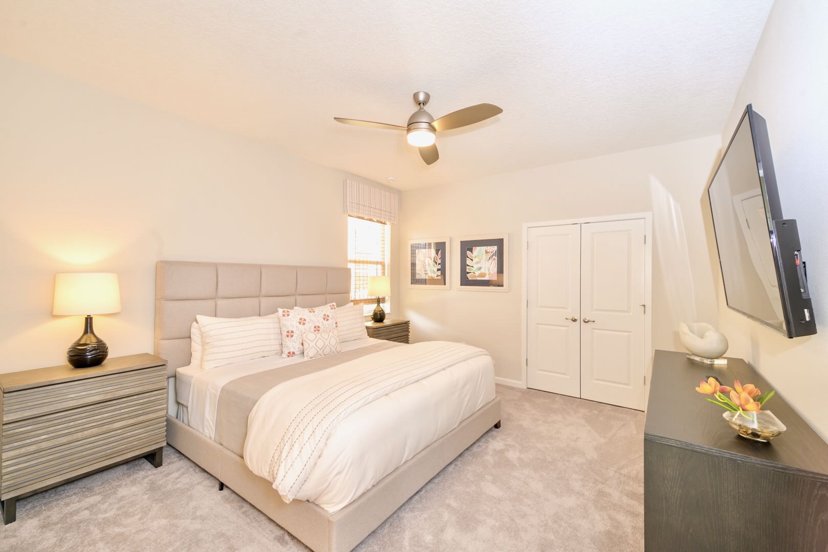 Bright bedroom with a large bed featuring a light upholstered headboard, white and patterned bedding, two nightstands with lamps, and a ceiling fan, all set on light gray carpet.