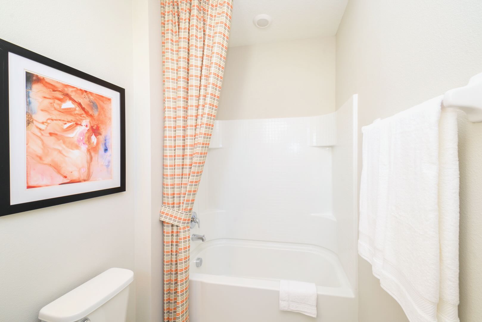 Compact bathroom with a white toilet and a white bathtub/shower combo. An orange and gray patterned shower curtain and a framed abstract art piece with orange tones add color to the light-colored space.