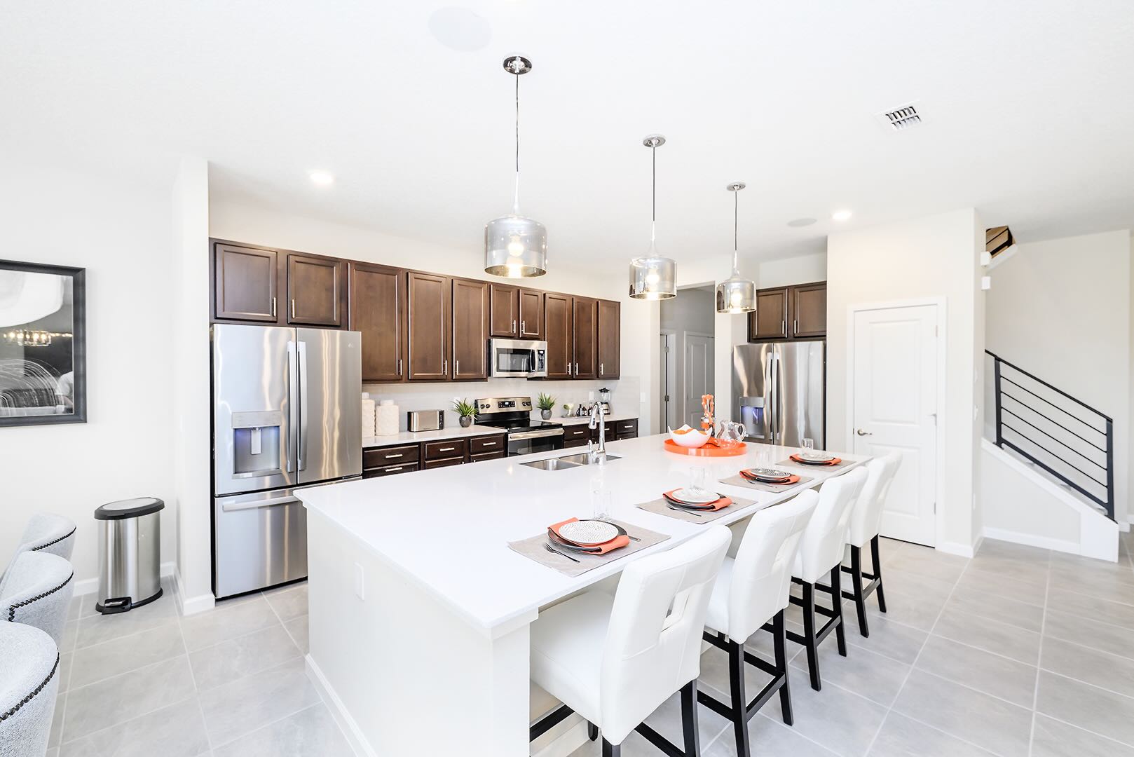 Spacious kitchen featuring a large white island with bar seating, dark wood cabinets, and stainless steel appliances including a double-door refrigerator. Light tile flooring and modern pendant lights complete the contemporary design.