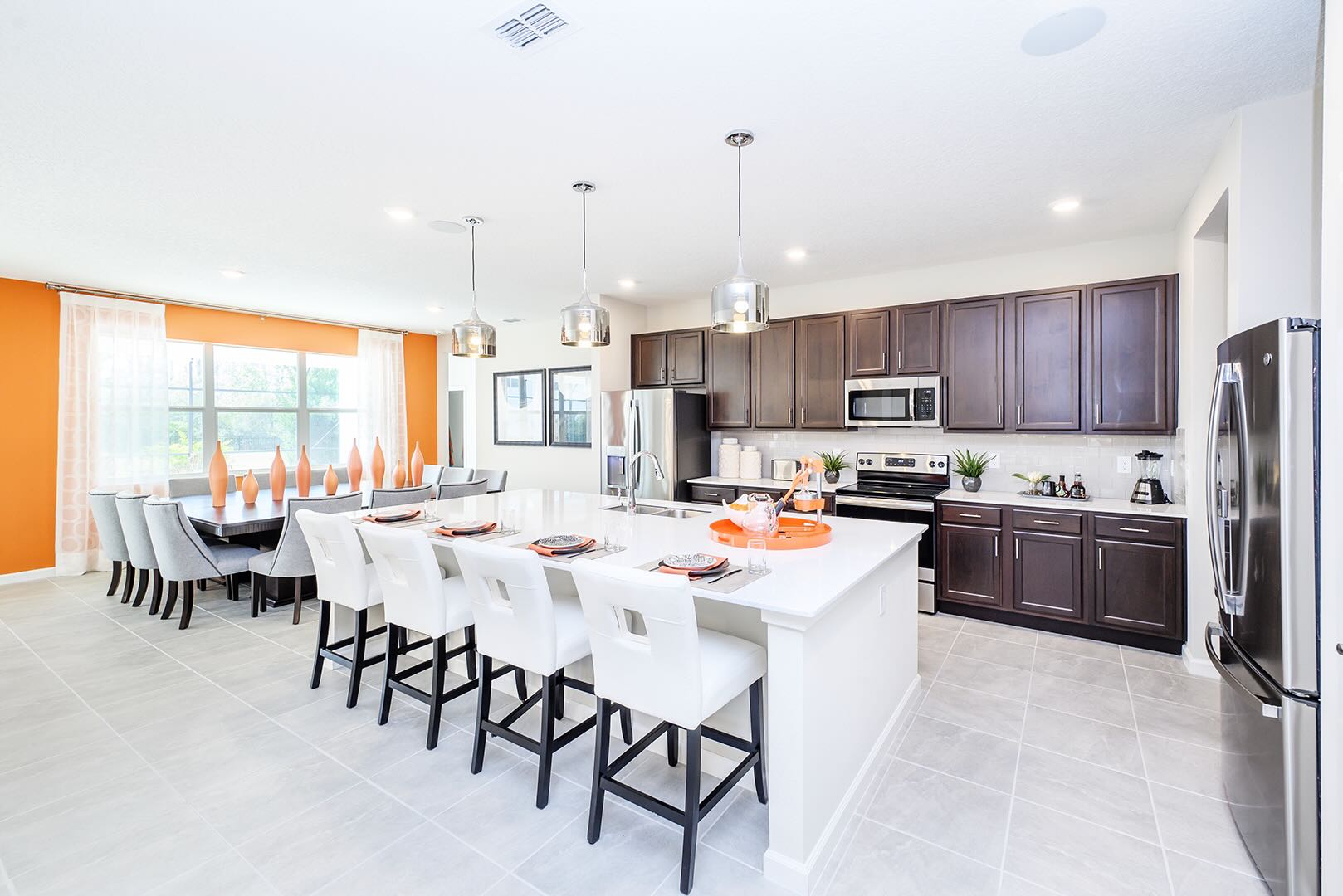 Modern kitchen with dark wood cabinetry, stainless steel appliances, and a large white island with seating for four. The adjacent dining area features a long table with numerous chairs, all under contemporary pendant lighting.