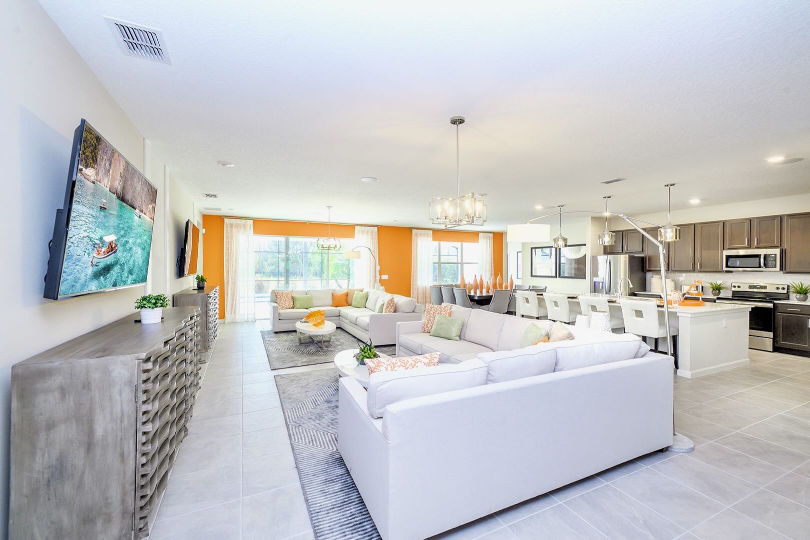 A spacious open-concept living area features two large L-shaped sofas with throw pillows, facing a wall-mounted TV above a long wooden console. Bright orange accent walls and modern chandeliers add pops of color and style, with a view into the kitchen.