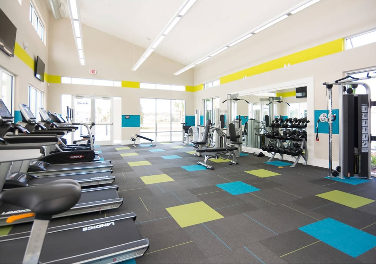 Bright and spacious fitness center with various exercise machines including treadmills, ellipticals, and weight benches, set on a colorful patterned carpet with large windows.