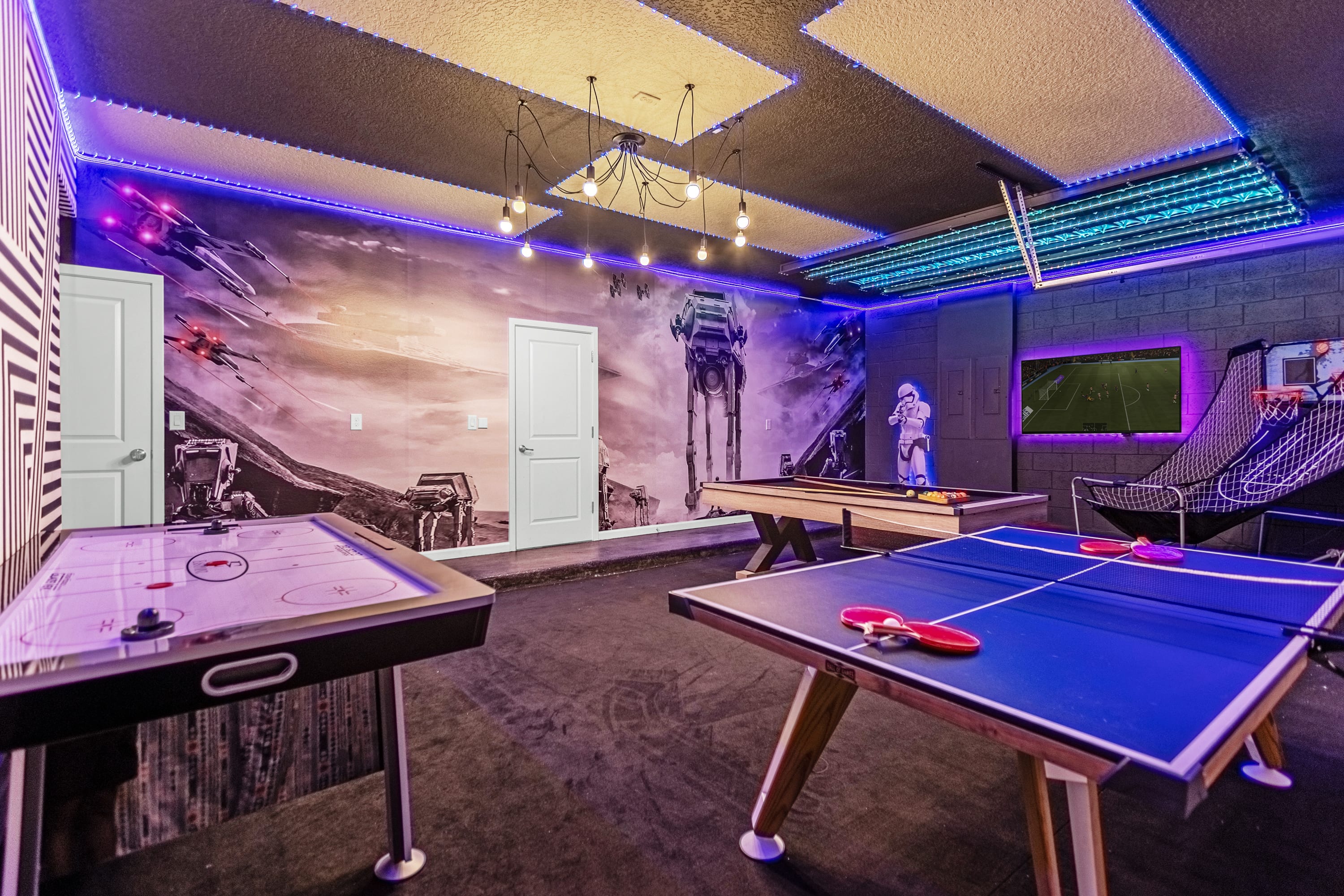 A vibrant game room with a Star Wars-themed mural covering one wall, featuring an air hockey table, a ping pong table, and a pool table. The ceiling has geometric acoustic panels and blue LED strip lighting, enhancing the playful atmosphere.