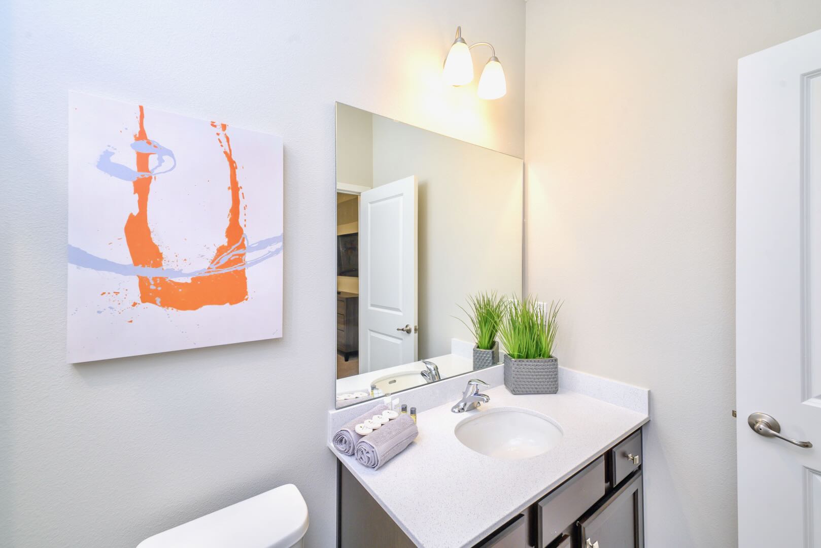 Modern bathroom with a white vanity, single sink, large mirror, and two-light fixture. An orange and blue abstract art piece hangs on the light gray wall, adding a pop of color.