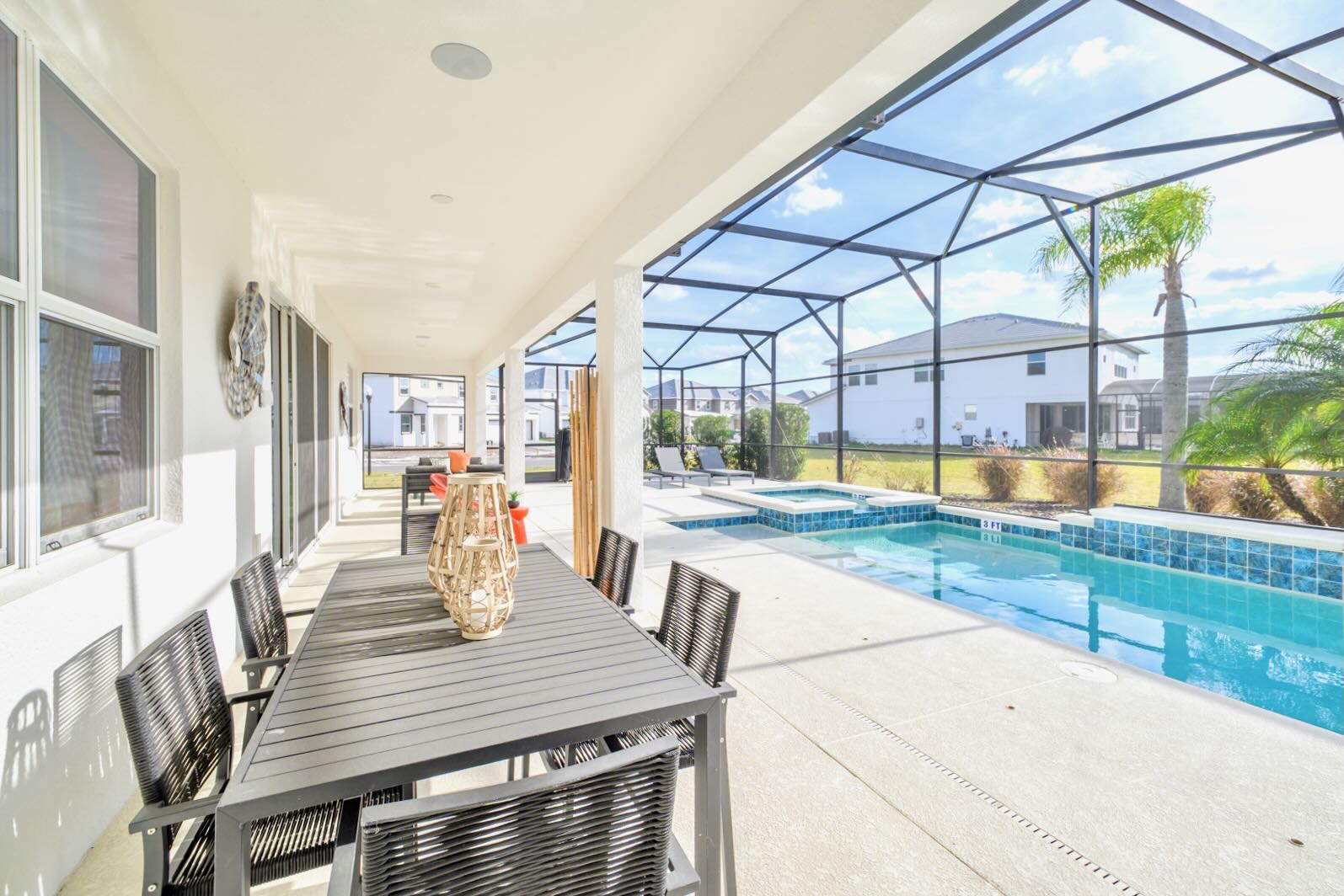 A spacious covered patio area adjacent to the house, featuring a large outdoor dining table with black woven chairs. The patio overlooks a screened-in pool and hot tub, with views of the sunny backyard and distant houses.